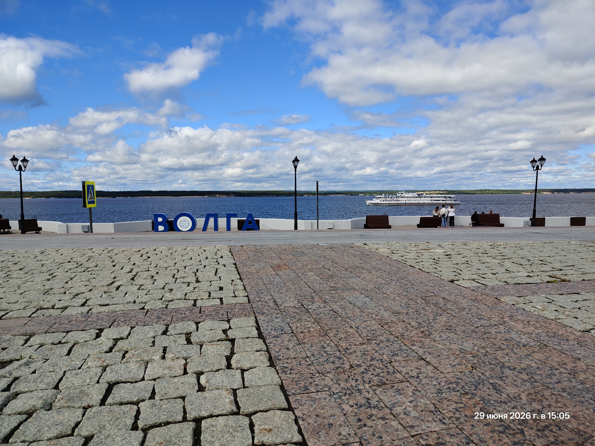 A wide shot captures a waterside promenade in an urban setting, likely on a sunny day with scattered clouds. The foreground is dominated by a textured cobblestone and granite pathway, with a subtle shift in material creating a visual division. In the middle ground, a blue body of water stretches to the horizon, with a large white passenger ship cruising on its surface. Along the waterfront, a white wall borders a paved area where several people are seated on benches, some standing and observing the boat. Prominently displayed on the waterfront is a large, blue, three-dimensional sign spelling out "ВОЛГА" (Volga). The background consists of a distant tree line and a bright blue sky filled with fluffy white clouds. A lamppost with two lights stands near the left side of the frame, and a pedestrian crossing sign is visible behind it. A timestamp in the lower right corner reads "29 июня 2026 г. в 15:05".