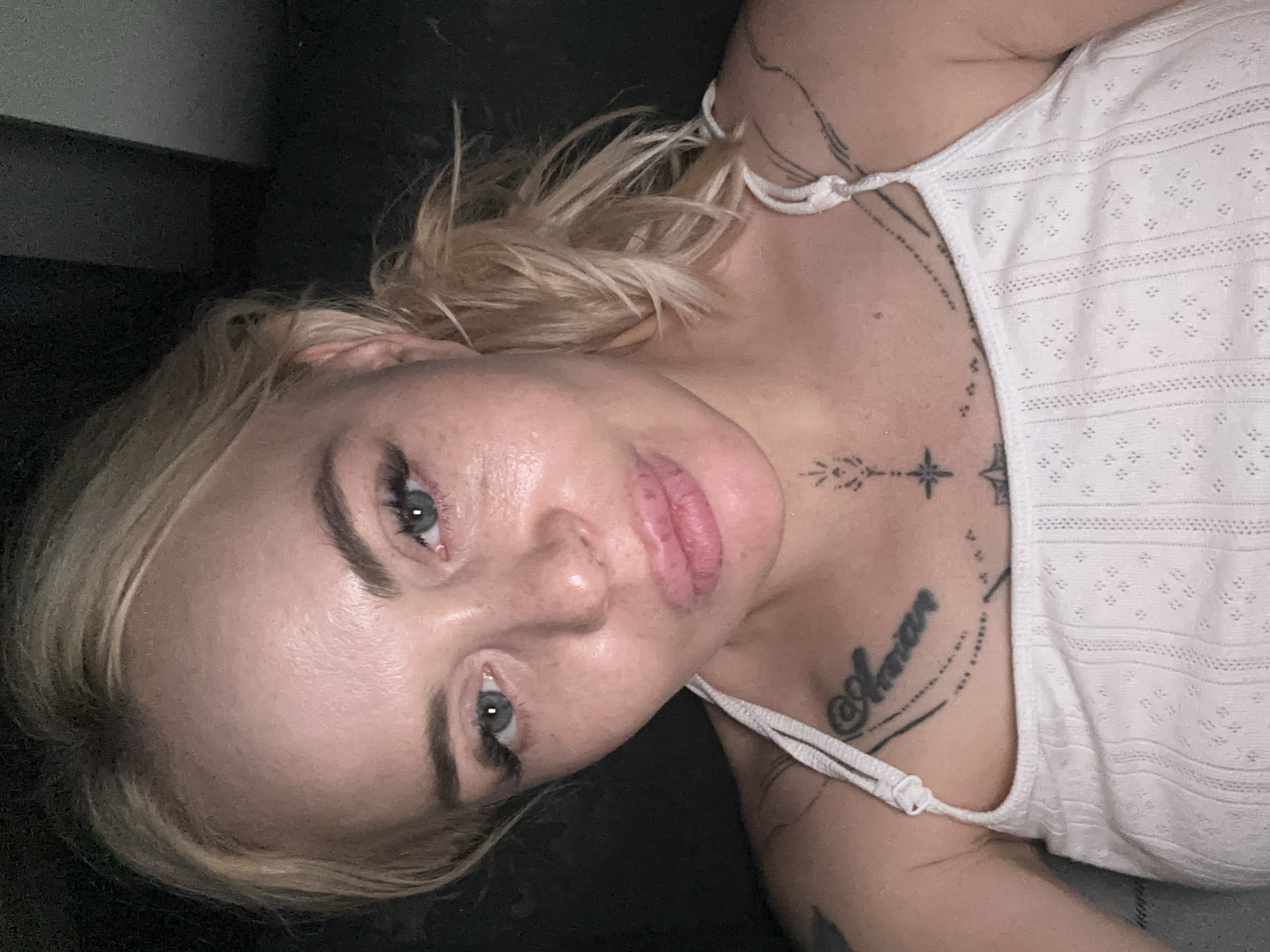 A close-up, eye-level indoor portrait captures a light-skinned individual with blonde hair tied back, posing with pursed lips and direct eye contact. The subject's light blue eyes feature prominent eyelashes and dark, shaped eyebrows. Her skin exhibits a slight sheen, particularly on the forehead and right cheek (viewer's left), where a scattering of faint freckles is also visible. She wears a white, textured camisole or tank top with thin straps, featuring a pattern of small vertical lines and diamond cutouts. Visible tattoos include script on the upper left chest (viewer's right) reading "Amore" or a similar word, with an accompanying trail of dots. Centered on the sternum is a symmetrical ornamental tattoo featuring a star-like element, radiating lines, and an arrow-like motif. Portions of additional tattoos are discernible on both shoulders and upper arms. The background is uniformly dark and indistinct, offering no specific details of the location in the city of Unknown, Unknown.