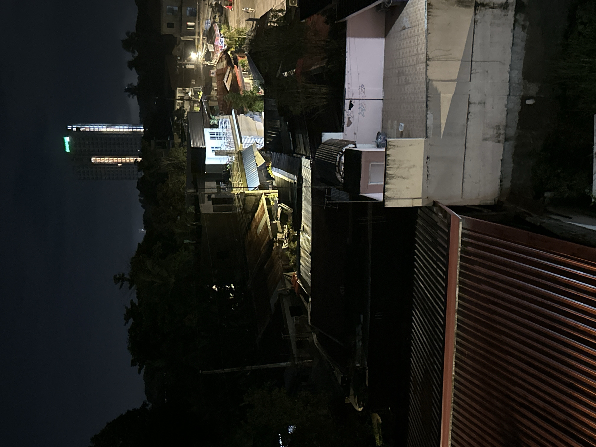 The image depicts a nighttime view of a city, likely in Davao City, Philippines, characterized by a mix of urban and residential structures. In the background, a prominent tall building, possibly a hotel or condominium, is illuminated with green and white lights. Below this, a densely packed area of smaller buildings with corrugated metal roofs is visible. Some of these structures appear to be houses or small businesses, with lights on in some of the windows. In the foreground, more rooftops are visible, some with red corrugated metal and others with lighter-colored tiling. The overall scene suggests a vibrant, bustling urban environment that continues into the night. The presence of dense housing and commercial buildings points to a residential or mixed-use district within the city. The lighting from the distant skyscraper and streetlights creates a contrast with the darker areas of the residential blocks.