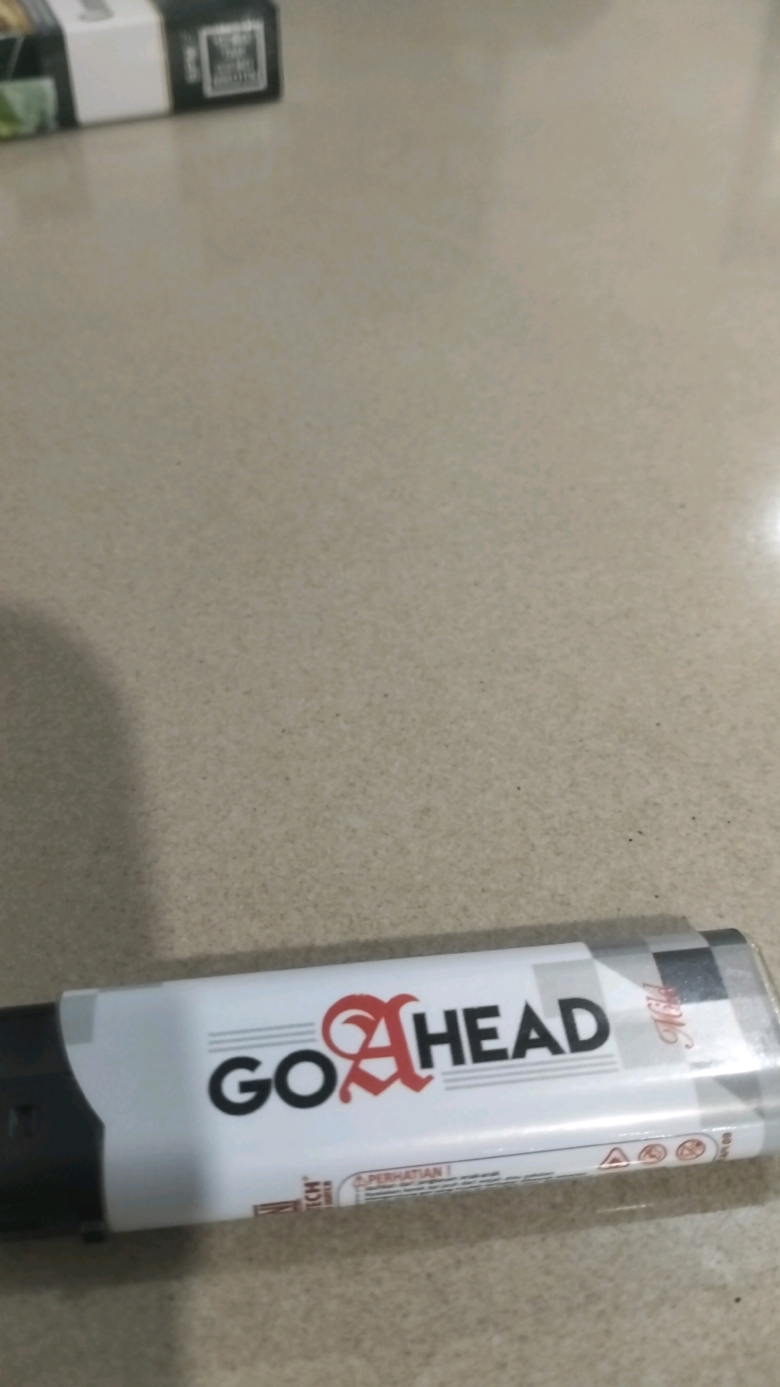 A white lighter with black and red lettering that reads "GO AHEAD" is lying on a speckled beige surface. The lighter has a black cap on the left side and a checkered pattern on the right. Below the "GO AHEAD" text, there is smaller text that appears to be warnings or instructions in Indonesian, including the word "PERHATIAN!" which means "ATTENTION!". The surface is likely a tabletop. The image is a close-up shot. There are no people visible in the image.