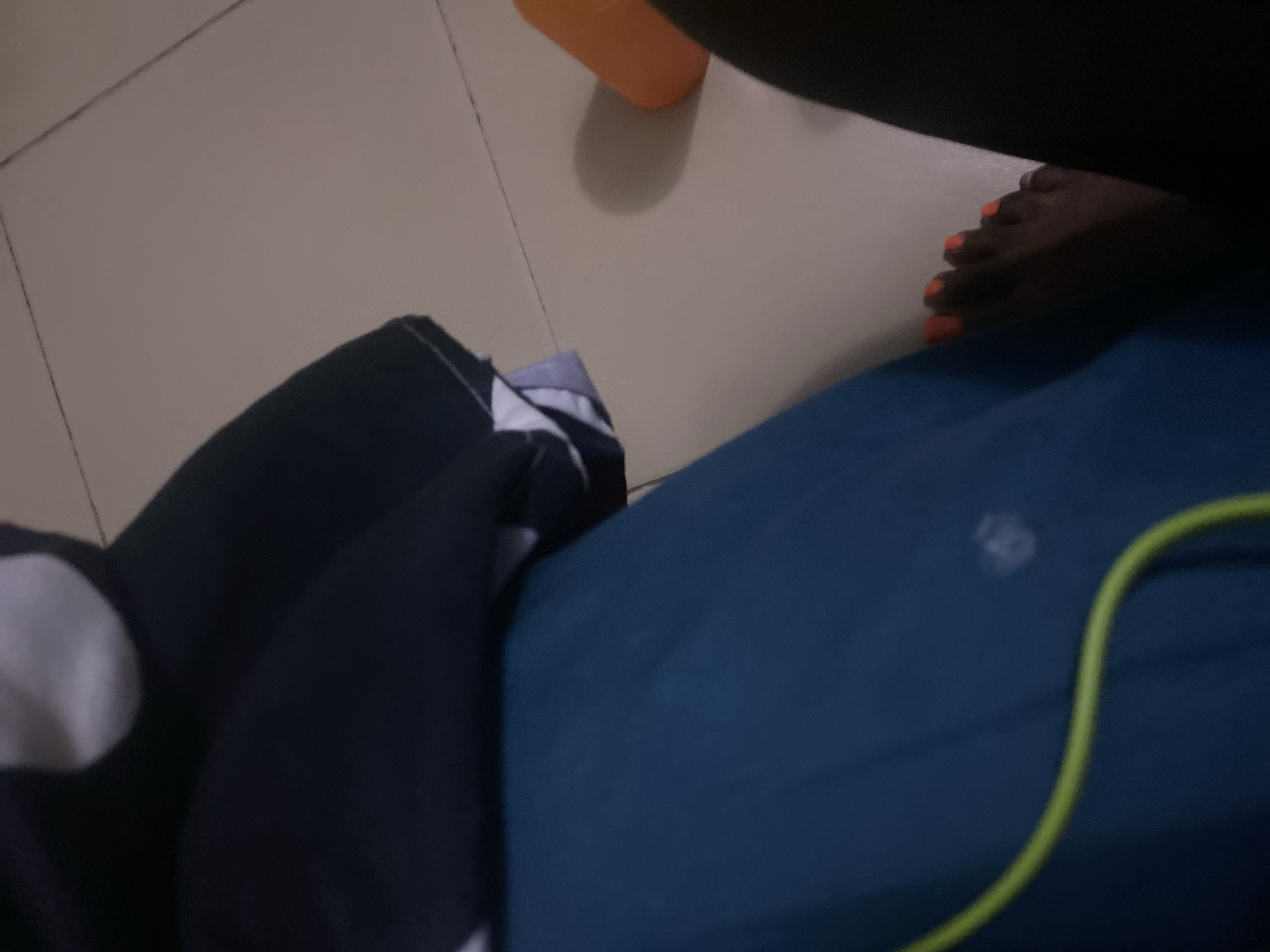 In a room with tiled flooring, a dark-skinned individual's feet are visible, adorned with bright orange nail polish. The feet are partially obscured by dark fabric on the right and a teal-colored garment on the left. A neon green cord, likely from headphones, is draped across the teal fabric. In the background, an orange plastic object casts a shadow on the floor. The scene appears to be indoors, in a domestic setting. The location is identified as Howayi, Nigeria.