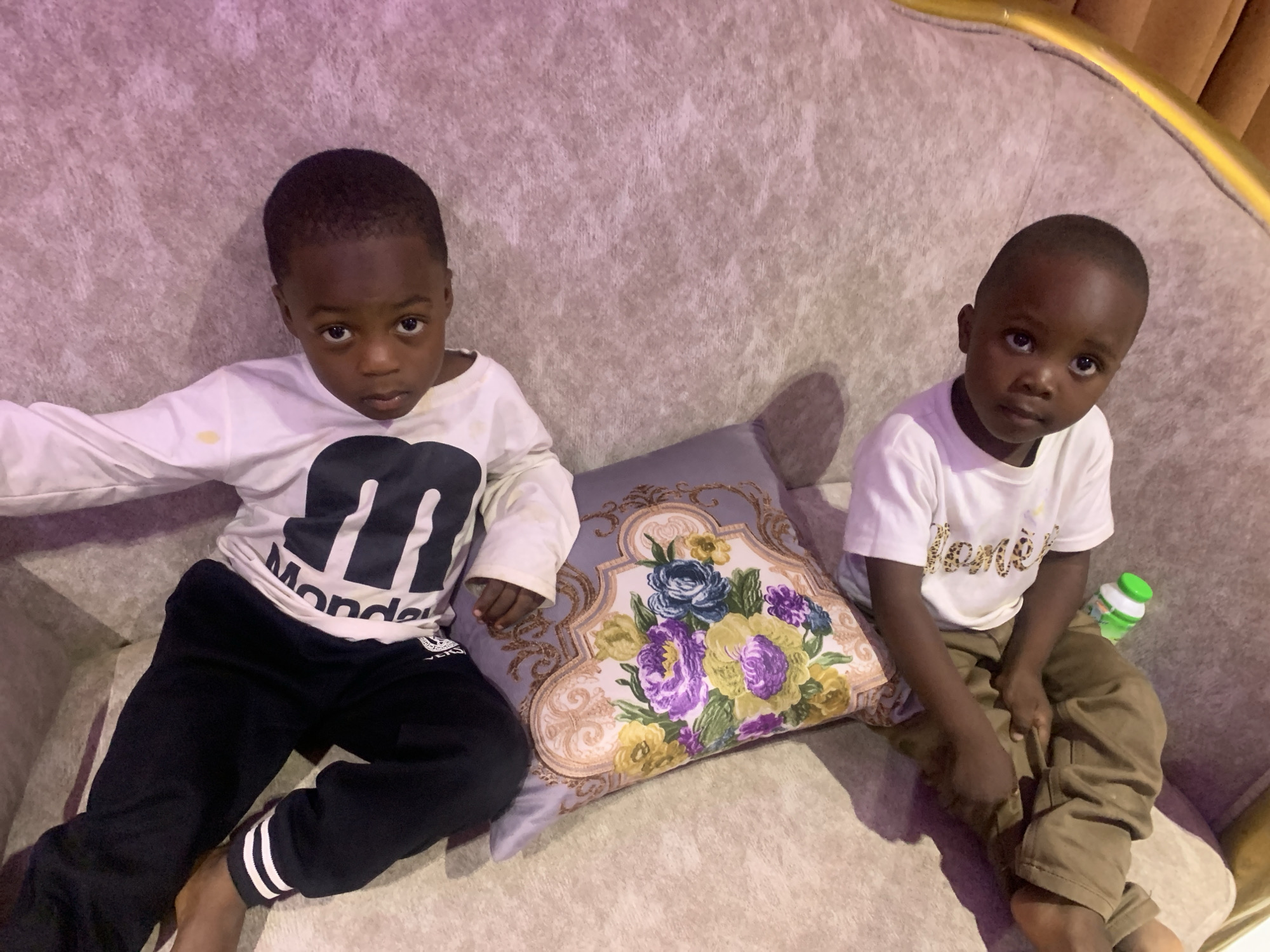 Two young, dark-skinned children are sitting on a light-colored, textured sofa in an indoor environment. The child on the left wears a long-sleeved, white shirt with black graphics and black pants with white stripes on the cuffs. The child on the right is wearing a short-sleeved, white shirt and khaki pants. A floral-patterned pillow is positioned between them. A bottle with a green lid is next to the child on the right. The location appears to be Jos, Nigeria, but cannot be definitively confirmed from the visual information.