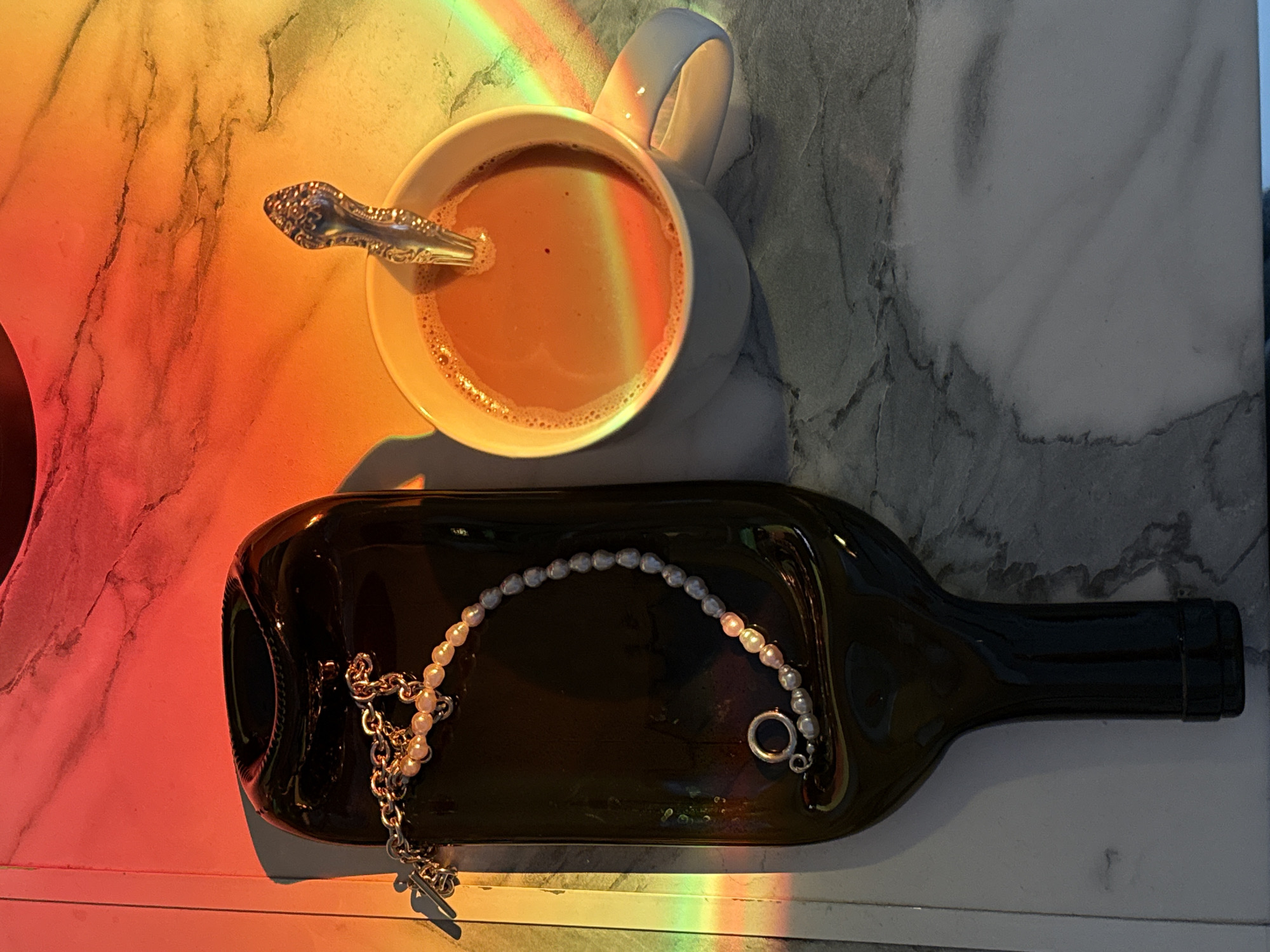The scene depicts an overhead view of two items resting on a light-colored, marble-patterned surface in Unknown, Unknown. A strong rainbow spectrum of light illuminates the upper portion of the scene, casting vivid red, orange, yellow, and green hues across the objects and surface. To the left, a dark, flattened glass bottle, possibly a wine bottle, serves as a tray. A bracelet featuring silver-colored chain links and a string of irregularly shaped pearls (some pinkish, some grayish) with a toggle clasp is draped across the bottle. To the right, a white ceramic mug, partially obscured by the rainbow light, contains a light brown beverage with a frothy top layer. A silver-colored spoon with an ornate handle rests vertically within the mug. No individuals are present.