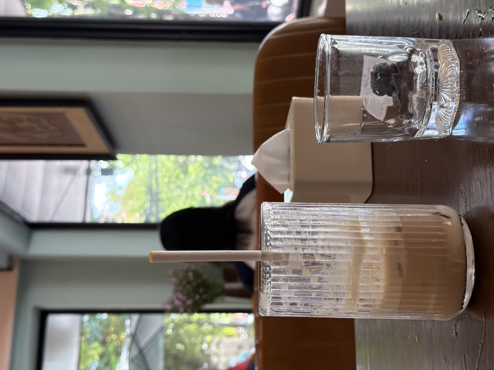 The image presents a close-up view of a tabletop scene, possibly in a café, located in Đà Nẵng, Vietnam. A glass of iced coffee with a straw is prominently displayed on the table. Adjacent to it is a clear glass with an indiscernible dark object submerged in a liquid within. A white tissue box sits between the two glasses. In the background, an out-of-focus view of a window reveals an outdoor setting with trees and a hint of urban structures, suggesting a location within the city. A person with dark hair can be seen through the window, contributing to the ambiance of a busy, urban location. The photograph is taken from a low angle, emphasizing the foreground objects. The colors are natural and warm, with good lighting. The background is heavily blurred, focusing attention on the drinks and the tissue box.