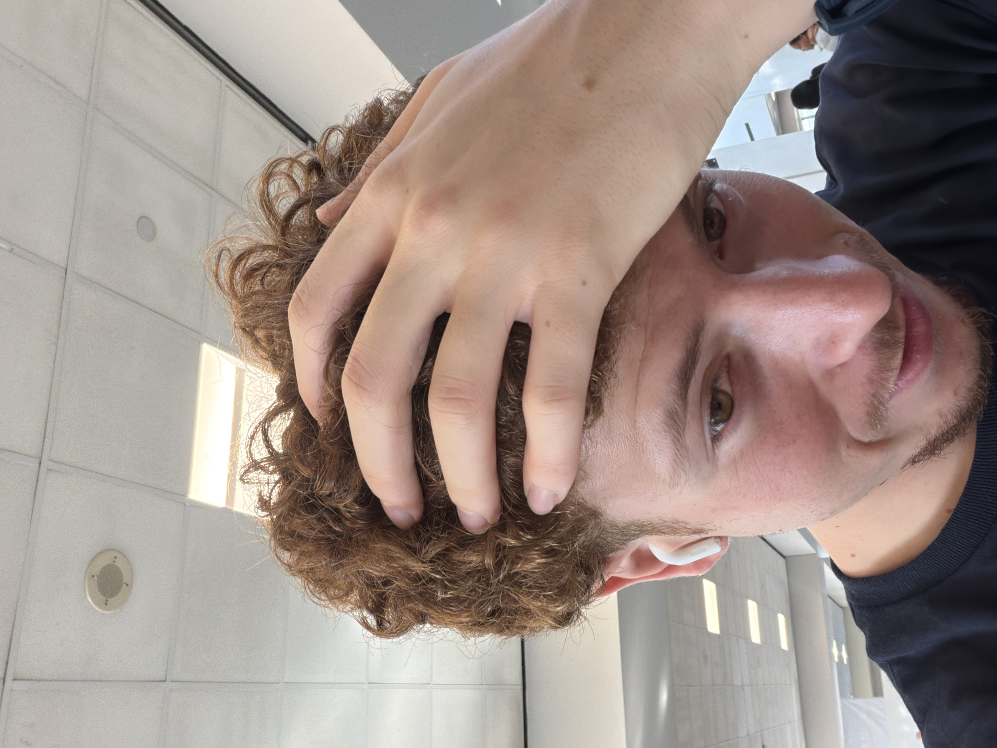 A man with brown curly hair, light skin, and a short beard is seen in a selfie taken at an angle looking up. His right hand is resting on top of his head. He is wearing a dark blue shirt and a white earbud is visible in his right ear. The background consists of a tiled wall and ceiling with bright, narrow vertical windows. The location is likely inside a building in an unknown area of Newark, United States of America.
