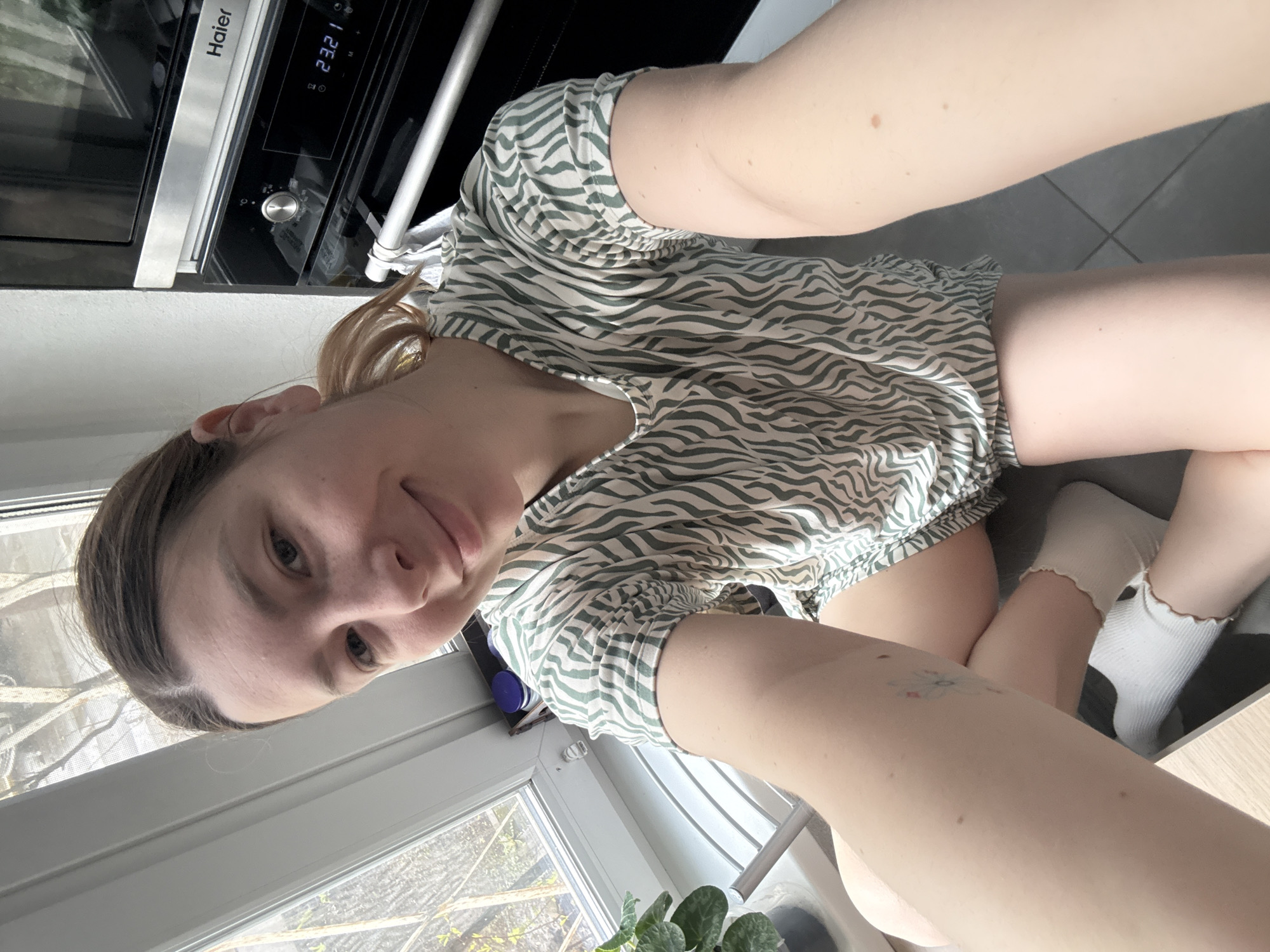 A woman with fair skin and light brown hair is kneeling indoors near a window. She is wearing a light green and white zebra-striped top and white socks with ruffled edges. Her left arm is extended towards the camera. A tattoo is visible on her right thigh.
Behind her are a kitchen counter and an oven. A potted plant sits on the windowsill. The view outside the window shows trees with sparse foliage.
The location is not identifiable as Moscow, Russia based on the provided image.