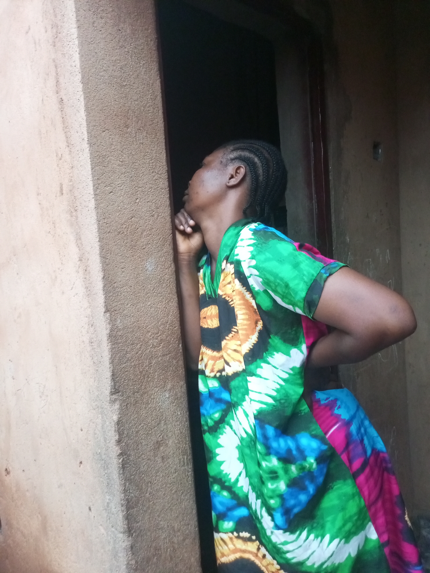 A Black woman with braided hair is standing in a doorway, looking outwards and to the left. She is wearing a brightly colored, flowing dress with a tie-dye pattern of green, blue, pink, yellow, and black. She is leaning against a textured, light brown wall on her left. The doorway behind her is dark and appears to lead into a building. The wall on her right is a darker, rougher texture. The scene is outdoors, and the lighting suggests it is daytime. There is no visible indication of the specific location in Tempure, Nigeria, beyond the general context of a domestic setting.