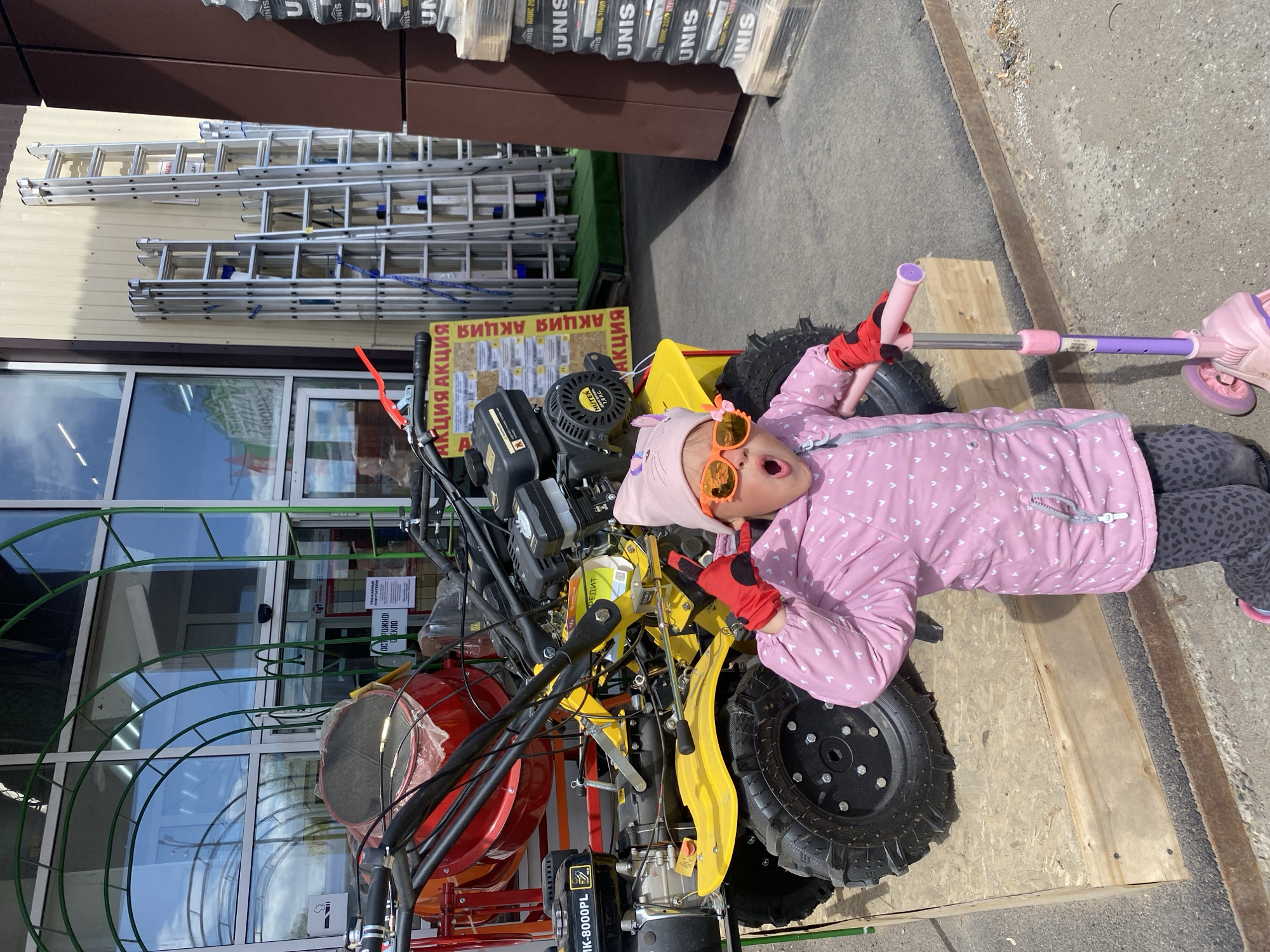 Here's a detailed description of the image:
**Scene:** The image appears to be taken outdoors, possibly in front of a retail store. The location is the city of Chaikovsky, Russia.
**People:** A young girl wearing a pink winter jacket, grey leopard print leggings, an orange and pink hat, and orange sunglasses is lying on top of a piece of agricultural machinery. She has red gloves on.
**Objects:**
* An agricultural machine with red and yellow components is visible. It seems to be a cultivator or similar garden tool.
* A pink and purple scooter is lying next to the girl.
* The retail store in the background appears to sell construction or home improvement items, as indicated by the rows of stacked products with "UNIS" branding.
* Ladders are stacked vertically behind the store.
* A sign with Russian text (presumably advertising an "AKCIA" or "Action" - Sale or promotion) is visible.
* A wooden board supports the machine.
**Actions:**
* The girl is lying down on the machinery and holding the scooter. She has her mouth open and appears to be either yelling or playfully surprised.
**Interactions:**
* The girl is interacting with the machinery and scooter, likely posing for the photo.
**Environment:**
* The scene is outdoors, with visible sunlight.
* The ground is paved.
* The background features a storefront and stacked merchandise.