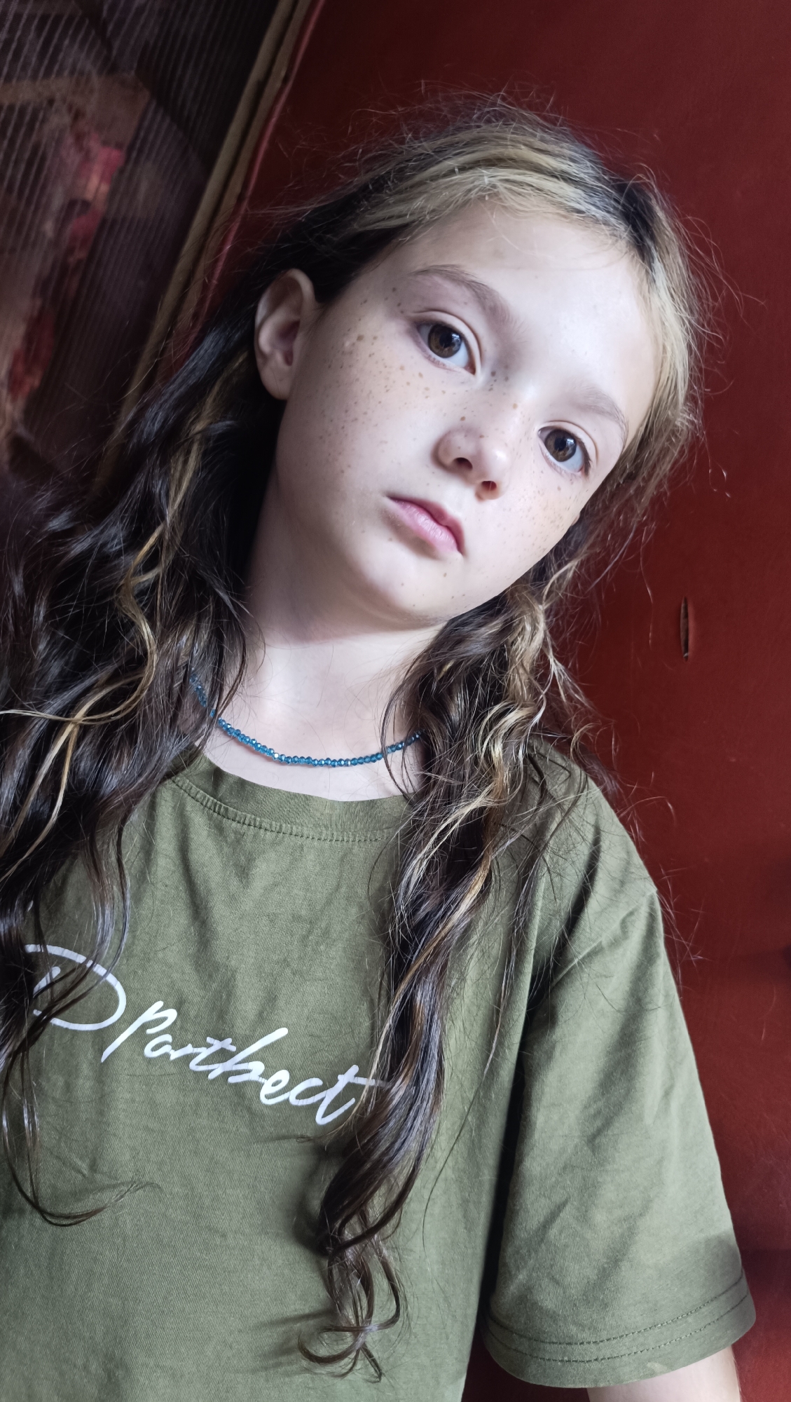 A young Caucasian girl with long, wavy brown hair, some streaks of blonde, and freckles across her nose and cheeks is looking at the camera. She is wearing a green t-shirt with white text that appears to say "Partheect" in a script font, and a thin blue beaded necklace. The background is a dark red, textured surface. The scene is indoors, likely a residential setting. The specific location in the city of Karsha, Russia is not identifiable from the provided image.