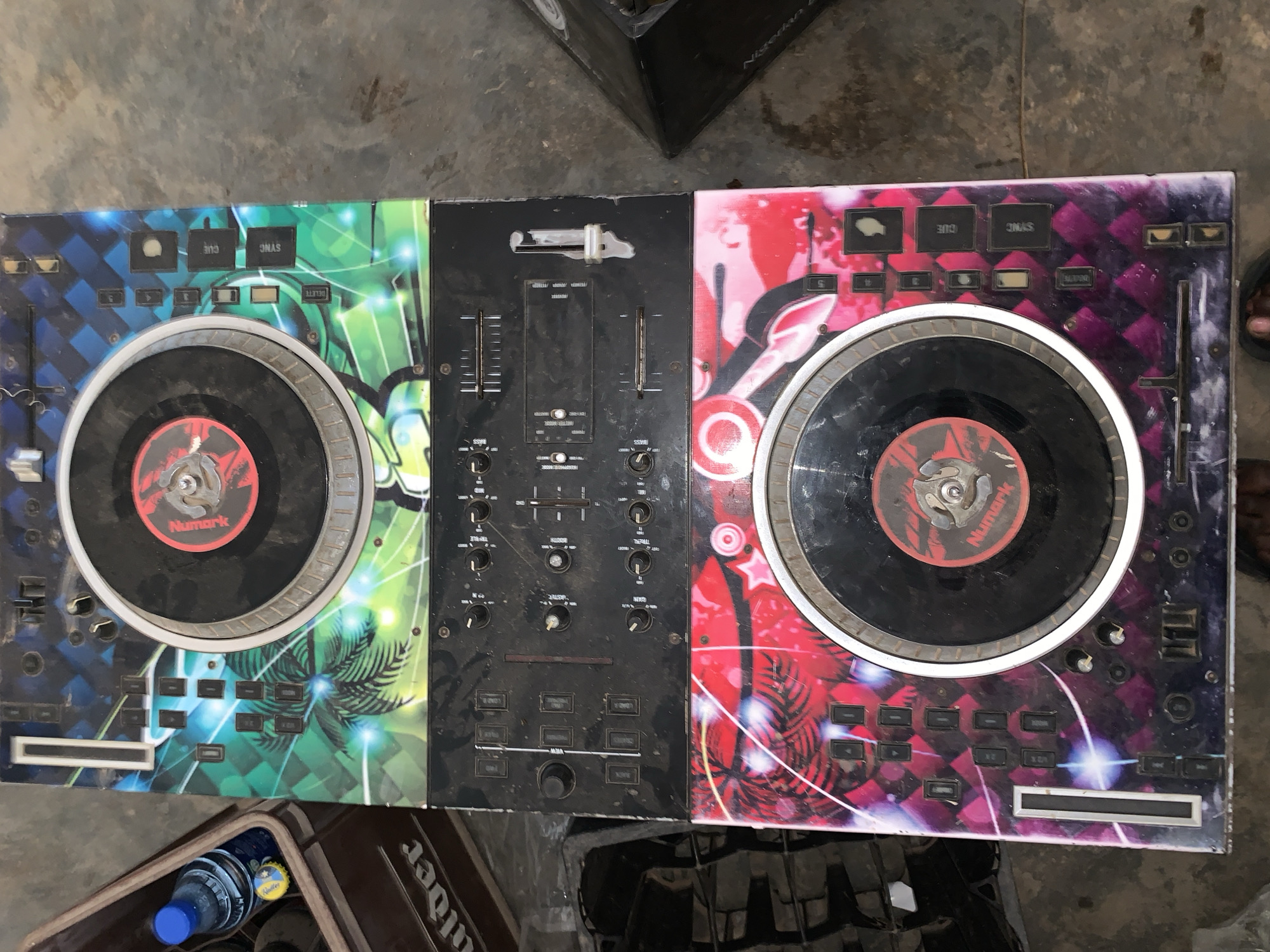 The image depicts a well-used Numark DJ controller set against a weathered concrete surface, likely in an outdoor or semi-outdoor location. The controller shows signs of significant wear and tear. The left deck is decorated with a blue, green, and black graphic, while the right deck has a red, purple, and black design. A mixer section with various knobs and faders sits between the two decks. Elements of a bottle and what appears to be a crate are visible at the bottom left of the image. The location in the city of New Stadium, Nigeria is not confirmed.