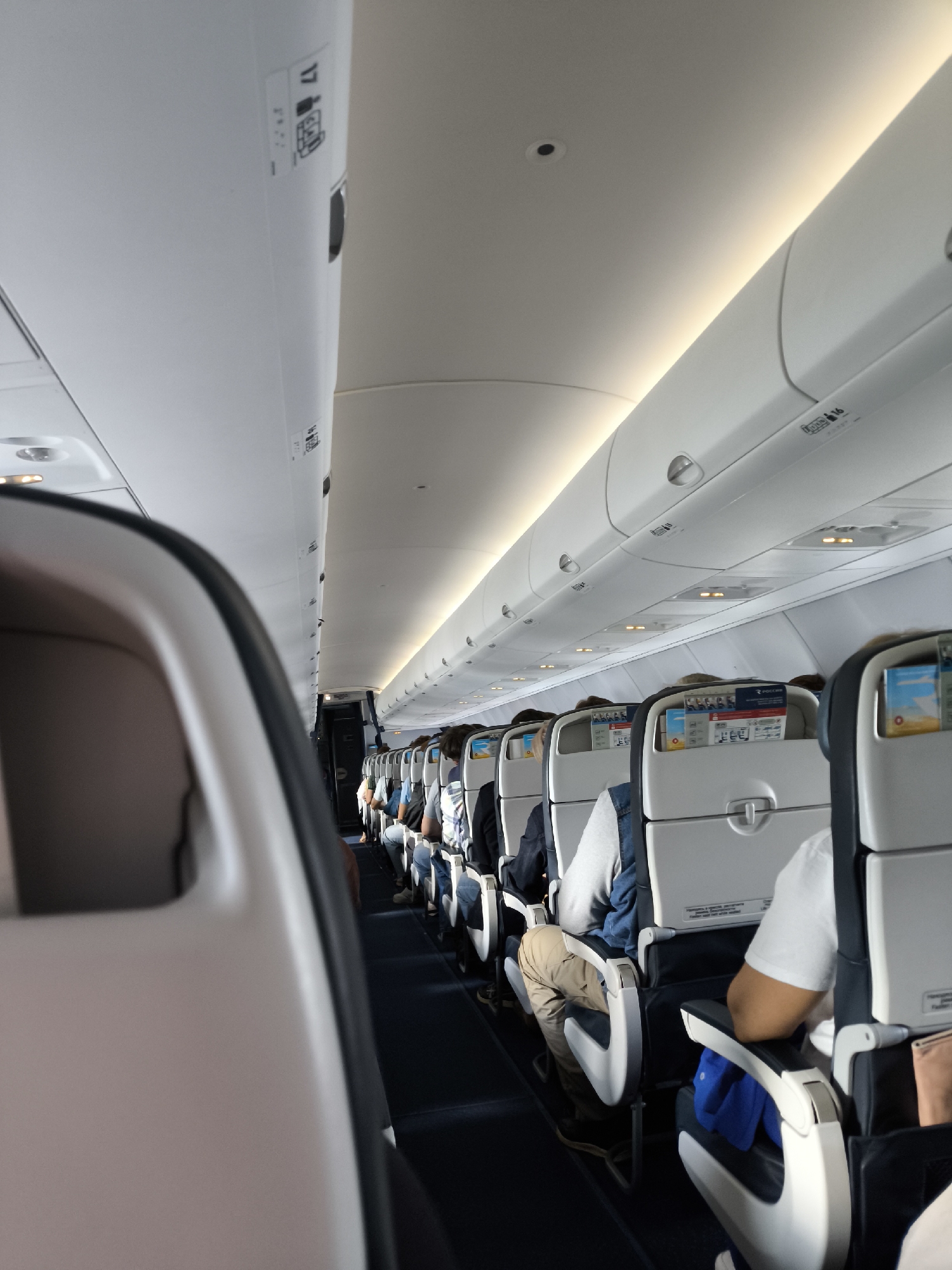 The image depicts the interior of a commercial airplane cabin, viewed from a slightly elevated position looking down an aisle from the front toward the rear. Rows of white-backed passenger seats with dark blue seat cushions and white armrests extend into the distance on both sides of a narrow, dark blue carpeted aisle. Multiple passengers are seated, primarily visible from the back, with their heads and shoulders, and some parts of their attire (such as khaki pants, blue jeans, and a white t-shirt) discernible.
Overhead compartments, uniformly white, line the cabin ceiling on both sides. These compartments feature latches and various small black symbols and text, including "OPEN" indicators and seat numbers like "17" and "16," along with luggage icons. The ceiling is curved and illuminated by continuous strip lighting, casting a warm glow. Additionally, individual overhead lights and air vents are visible above each seating section. In the seat-back pockets, safety instruction cards and other reading materials are visible, with one clearly displaying the word "РОССИЯ" (Russia). The foreground shows the rounded edge of a seat in soft focus, indicating the camera's position within a seat. The cabin environment appears well-lit and orderly.