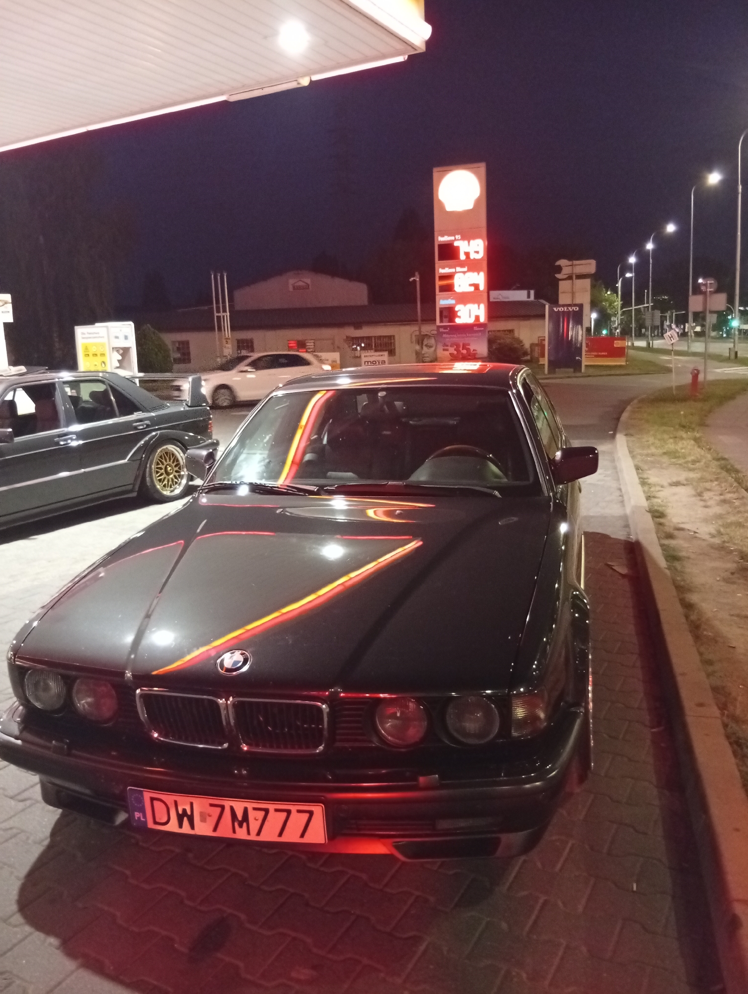 This is a night-time, medium-shot, eye-level, outdoor photograph of a dark colored BMW E32 7 Series sedan parked at a Shell gas station. The car is facing the camera and is mostly in focus. The license plate reads "DW-7M777". The car has red and yellow stripes on the hood that form a "V" shape. A second dark colored car, possibly a Mercedes-Benz, is parked behind and to the left of the BMW. The Shell gas station has a lit up sign displaying fuel prices: "Premium 95 7.49", "Premium Diesel 8.24", and "Gaz 3.04". A white Volvo sign is visible in the background. The image appears to be taken in Wrocław, Poland, as indicated by the license plate format and the gas station signage. The environment is dimly lit, with the car's headlights and gas station lights illuminating the scene. The ground is paved with cobblestones. The sky is dark and appears to be clear.