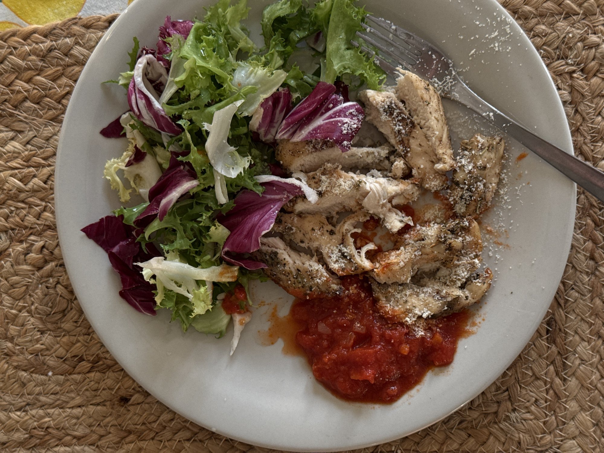 An overhead, close-up shot displays a white round plate filled with a meal, resting on a light brown, woven placemat. A portion of a yellow and white patterned tablecloth is visible in the top left corner.
The plate features several components:
On the right, slices of cooked poultry, likely chicken, are seasoned with herbs and generously topped with grated white cheese, resembling Parmesan. The chicken appears to be pan-fried or baked.
To the left of the chicken, a fresh side salad is arranged, composed of mixed greens including bright green lettuce and darker red radicchio leaves. Some grated cheese is also visible on the salad.
At the bottom of the plate, a portion of chunky red sauce, consistent with a tomato or marinara sauce, accompanies the chicken, with some liquid seeping onto the plate.
A silver fork is partially visible on the upper right side of the plate, suggesting the meal is ready to be eaten or is already in progress. The lighting is bright and even, indicating a daytime setting. The composition focuses entirely on the plate and its immediate surroundings. There is no visible text in the image.
