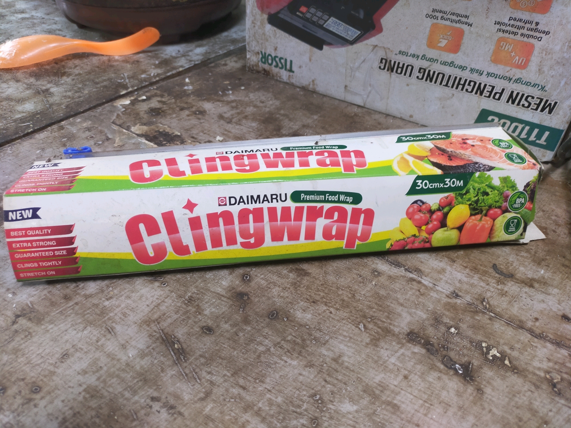 The image depicts a close-up, eye-level view of a roll of "Clingwrap Premium Food Wrap" by Daimaru, lying on a weathered, textured wooden surface. The packaging is predominantly white with vibrant red and green lettering, featuring images of salmon, lemons, and various fruits and vegetables. The roll is oriented horizontally across the frame. To the left of the clingwrap, a bright orange plastic spoon is visible, partially obscuring a brown, circular object. In the background, a portion of a cardboard box with Indonesian text is visible, suggesting a retail or storage environment. The wooden surface exhibits signs of wear, with visible grain, scratches, and scattered debris. The lighting is natural and diffused, casting soft shadows. The scene is devoid of human presence, indicating a still-life composition focused on the product and its immediate surroundings. The location is identified as Mojodanu, Indonesia, implying this is likely a scene from a local market or household setting.