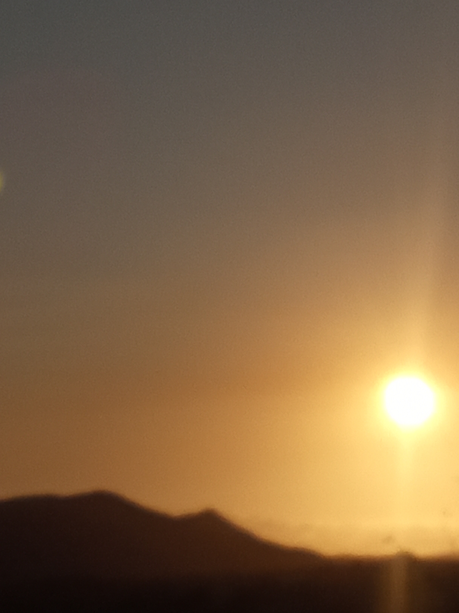 This image captures a landscape view at either sunrise or sunset over Juan Griego, Venezuela. The sky occupies the majority of the frame, displaying a gradient of colors from a dark gray-blue in the upper left transitioning to a vibrant orange and yellow on the right. A prominent, intensely bright, circular white-yellow sun is visible in the lower right quadrant, emitting strong illumination and lens flare effects. A dark, indistinct mountain or hill range, silhouetted against the brighter horizon, extends across the lower third of the image. No individuals, man-made structures, or specific actions are discernible within the scene.