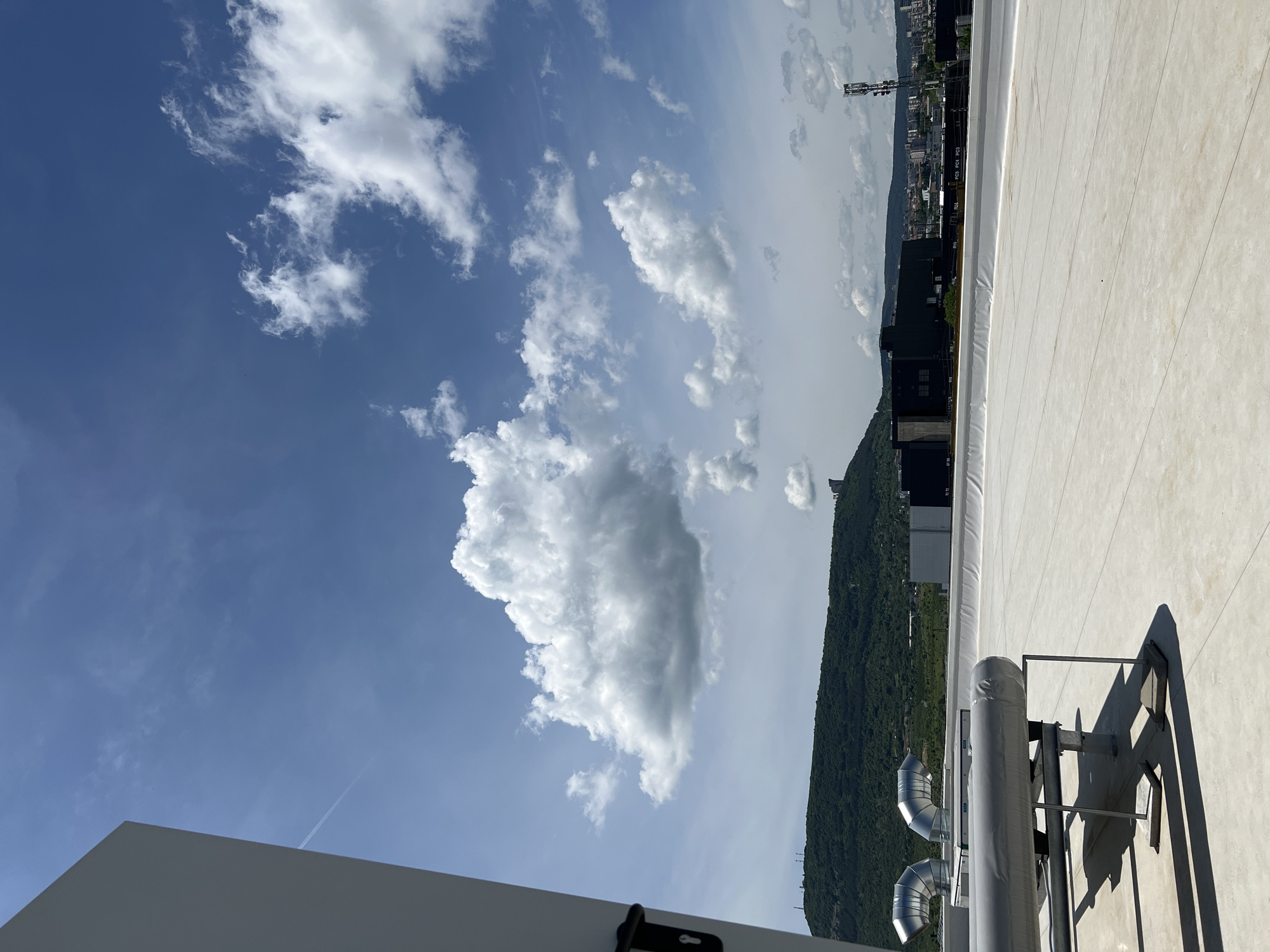 The image presents a bright outdoor scene from an elevated vantage point, likely a rooftop, under a clear blue sky. The composition features a foreground of architectural elements and equipment, a prominent sky with clouds, and a distant landscape of mountains and urban development.
In the immediate foreground, a large, light-colored flat surface, consistent with a rooftop, spans the right side of the image. On this surface, industrial equipment is visible, including silver-colored, insulated pipes or ducts supported by a metal framework, all casting distinct, sharp shadows indicative of bright sunlight. A smaller, curved silver duct is also seen nearby. Towards the bottom left, a section of a white or light gray building structure is present, featuring a black handle or latch.
The sky occupies a large portion of the upper and middle sections of the image. It is a vibrant blue with scattered white cumulus clouds of varying sizes and shapes, from large, fluffy masses to more delicate, wispy formations. A faint, thin contrail from an aircraft is noticeable in the upper left corner of the sky.
Stretching across the mid-ground and into the background is a verdant, forested mountain or large hill. Beyond this natural barrier, in the far distance, a cityscape can be observed, comprising numerous buildings and structures under a slightly hazy sky. On a dark rectangular object within this distant urban setting, the white text "PC1 PC2 PC3 PC4" is faintly visible.
The scene depicts a sunny day with fair weather, evidenced by the bright light and clear shadows. No human activity or specific event is taking place; it's a static landscape and architectural view.