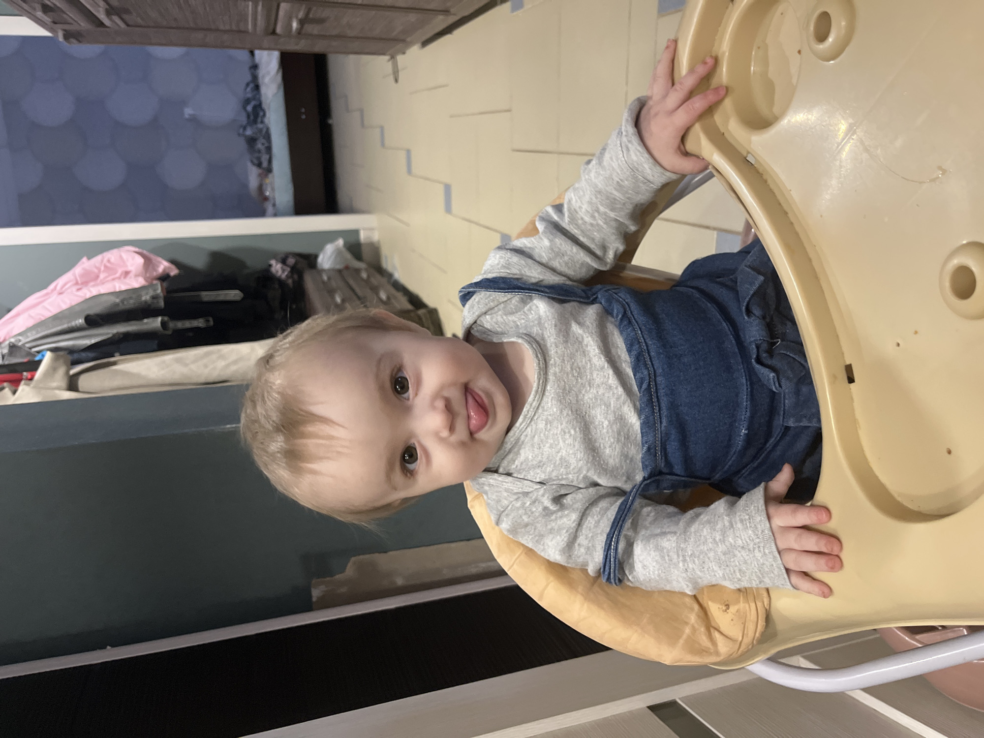 A young child with light-colored hair and dark eyes is depicted sitting in a beige plastic high chair, facing forward, with their tongue extended. The child is wearing a grey long-sleeved bodysuit beneath blue denim overalls. Their left hand rests on the high chair's tray, which appears empty. The background consists of an indoor domestic setting in Chaikovsky, Russia. To the immediate left of the child, a dark wall is visible, along with hanging clothes, including a light pink garment and darker jackets. A light-colored cabinet or dresser with multiple drawers is partially visible below the hanging clothes. To the right, a doorway leads into another room featuring blue patterned wallpaper with circular motifs and a bed with a light blue mattress and patterned bedding. A dark wooden cabinet or wardrobe stands on the far right. The floor is covered with light-colored tiles accented with blue lines forming a zigzag pattern.