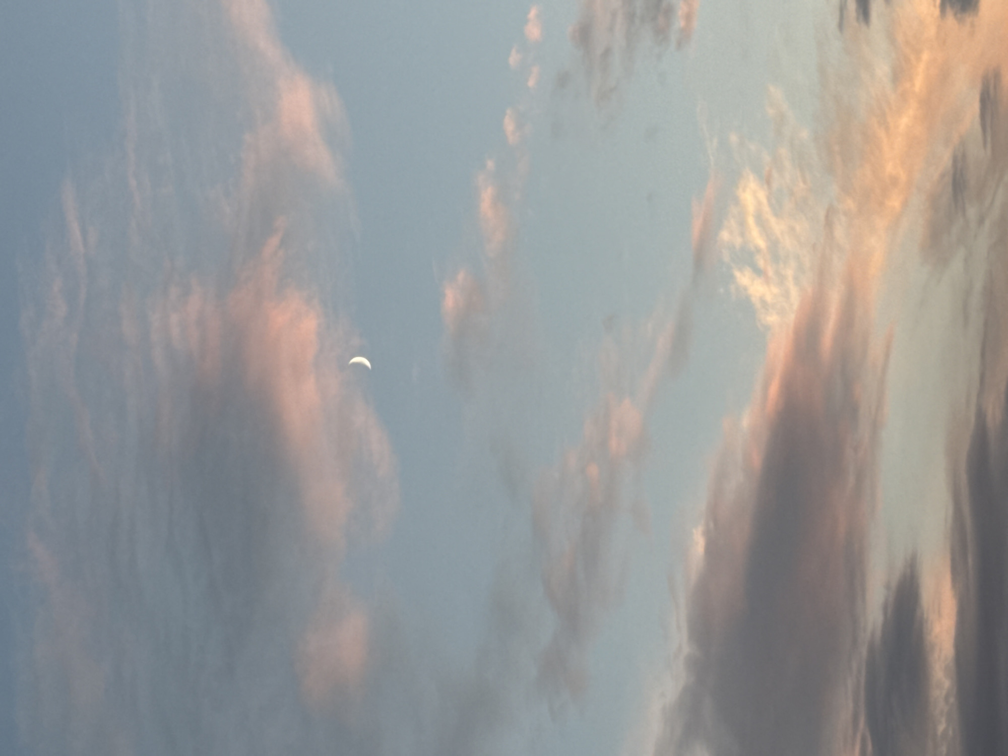 An image captures a serene sky with a crescent moon hanging in the upper quadrant. The sky is painted with hues of soft blue, transitioning into delicate shades of pink and orange from the setting or rising sun, creating a beautiful gradient. Wispy clouds, tinged with the same warm colors, drift across the canvas of the sky, some appearing more defined and others more ethereal. The overall impression is one of tranquility and natural beauty in the vast expanse of the sky.