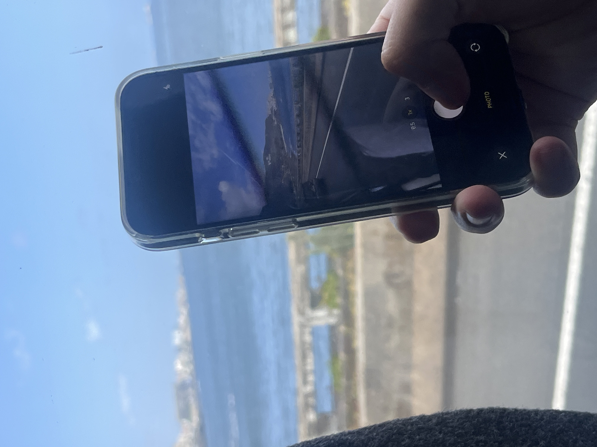 The image captures a person's hand holding a smartphone horizontally, with its camera application open, suggesting the act of taking a picture. The phone is enclosed in a clear protective case.
The person's thumb is positioned near the screen, on the right side of the phone. The smartphone's display shows a camera interface, indicating "PHOTO" mode. Visible on the screen are zoom options "0.5", "1x", and "3", as well as a circular white shutter button, a rotate camera icon, and an "X" button. A small sensor or dot is visible on the phone's top bezel. The screen also partially reflects the surrounding environment.
The scene outside the window, visible around and reflected on the phone, consists of a vast blue sky with minimal cloud cover and a wide expanse of blue water, indicative of a sea or ocean. In the distance, a mountainous or hilly coastline with visible structures or buildings is discernible, stretching along the horizon. The view suggests a bright, clear day, likely sunny. The immediate foreground at the bottom is a dark, textured material, possibly upholstery from a vehicle or furniture, implying the photo is taken from indoors looking out. The coastal landscape with bright skies and clear waters is consistent with the location context of Telde, Spain.