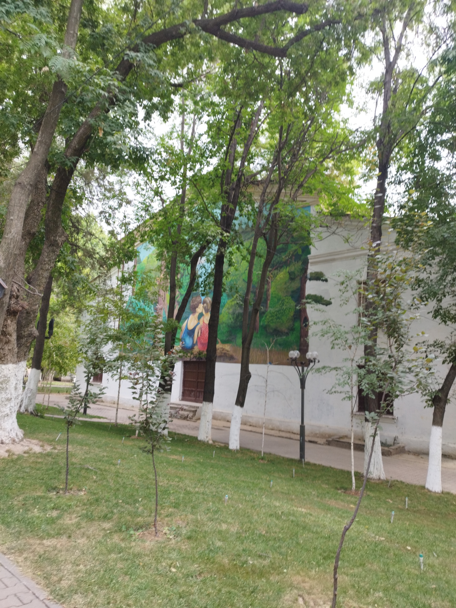 The image depicts a mural on the side of a white building, featuring a landscape scene with two figures in the foreground. The mural shows a forest with tall trees and green foliage. The two figures appear to be a couple kissing, with the person on the left wearing a blue top and the person on the right wearing a red top. The building has a brown door. In front of the building, there is a paved walkway and a grassy area with several young trees. Mature trees with green leaves are positioned in front of the building, partially obscuring the mural and the building itself. The base of some of the mature trees is painted white. A lamppost with three lights is visible in front of the building, between the trees. The overall setting suggests an outdoor public space, possibly a park or a courtyard. This mural is located on the side of the Shymkent Regional Philharmonic Hall in Shymkent, Kazakhstan.