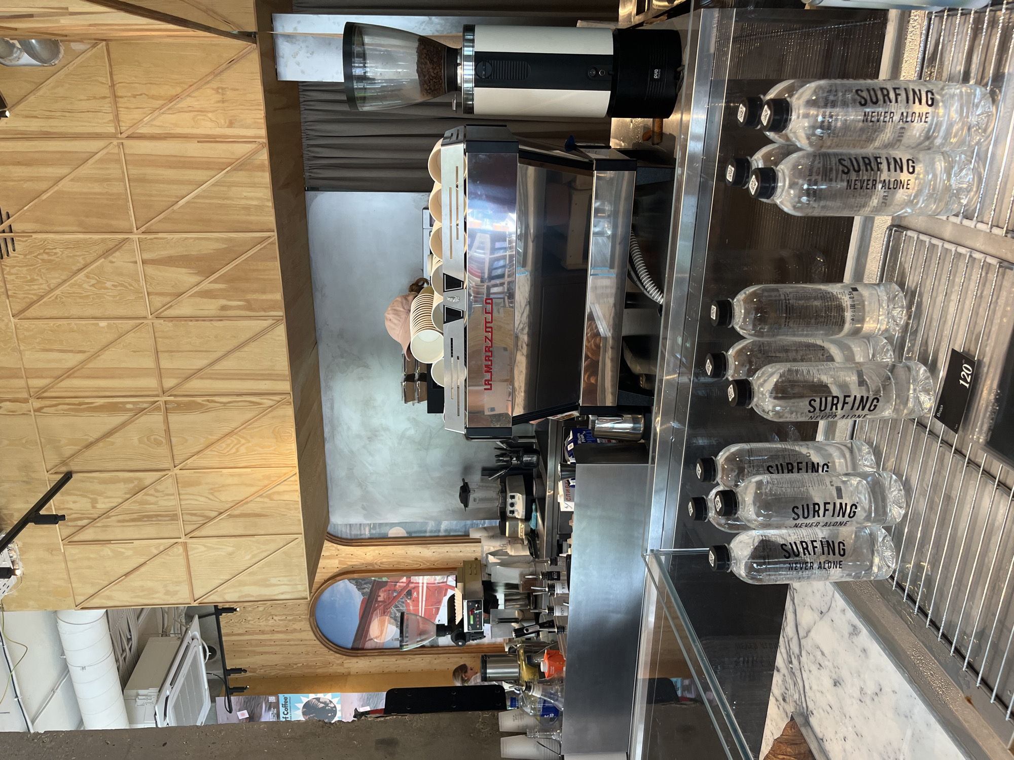 This image depicts the interior of a coffee shop, likely located in Estosadok, Russia, as suggested by the presence of a price tag with the Russian word "Вода" (water) and the number "120". The scene is dominated by a polished stainless steel counter and a large espresso machine. Behind the counter, wooden panels with a geometric design are visible on the wall, along with a large arched mural.
In the foreground, several clear plastic bottles of water with black caps are arranged on a metal grate. The labels on these bottles clearly read "SURFING NEVER ALONE". A croissant is partially visible on a tray to the left of the water bottles.
Further back, on the counter, the espresso machine is prominent, with a coffee grinder to its right. Various cups and other coffee-making accessories are visible on top of and around the machine. A person's head and shoulders are visible in the background, suggesting an employee is working behind the counter. The overall environment appears clean and modern, with elements of natural wood and stainless steel contributing to a contemporary aesthetic. The lighting suggests it is daytime.