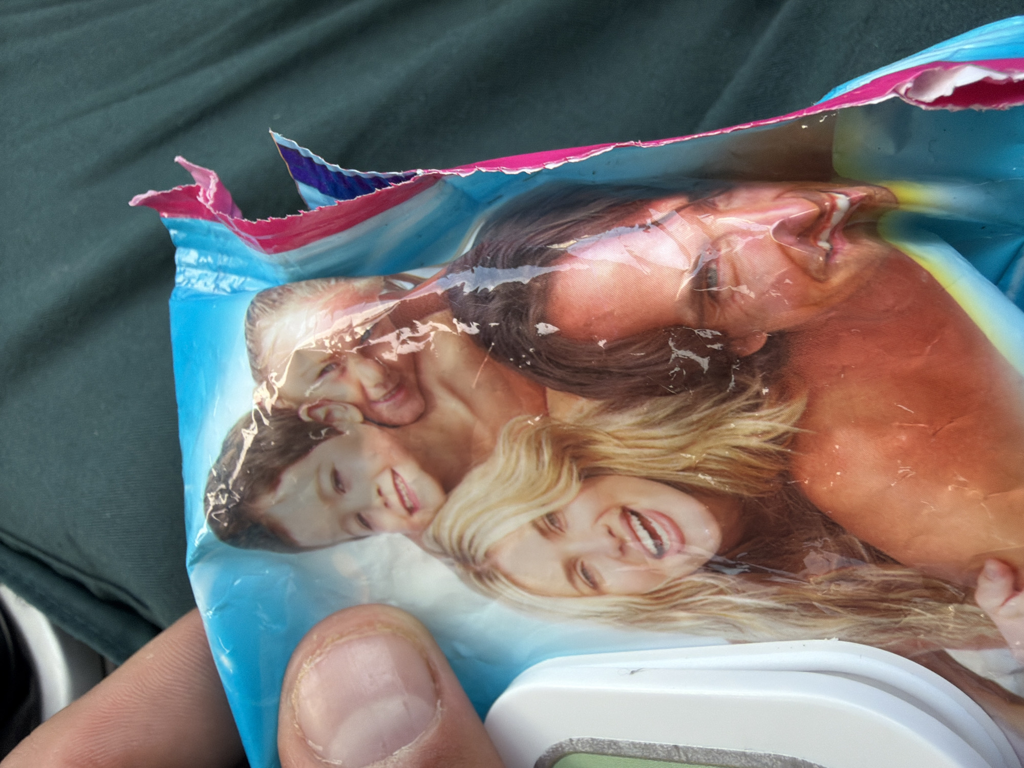 An adult's left hand, characterized by light skin and short, natural fingernails, holds a partially torn plastic package. The package is primarily light blue and features a printed collage of four smiling Caucasian individuals: a young boy with light brown hair at the top, an adult blonde woman with wavy hair and an open-mouthed smile (winking with her left eye) on the lower-left, and an adult man with dark wavy hair and a broad, open-mouthed smile on the lower-right. A fourth adult figure is partially obscured behind the boy in the upper-right. The plastic surface of the package is crinkled, and its right edge is torn, revealing layered pink and dark blue material. The immediate background behind the package is dark teal or green folded fabric, indicative of clothing, possibly a person's lap or shirt. A white plastic or ceramic object with a thin green stripe is partially visible in the lower-left corner of the frame. The image does not contain any visual indicators to confirm or deny the requested location of Rezovo, Bulgaria.