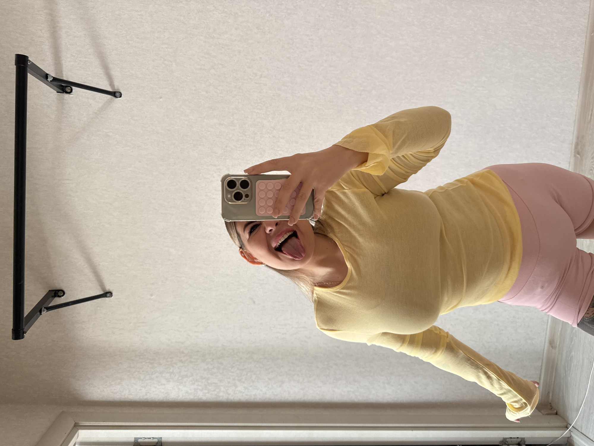 A woman wearing a yellow top and pink shorts is taking a selfie in what appears to be a room. The camera is held in her right hand, and she is sticking her tongue out towards the camera. On the wall is a black metal structure, possibly a clothing rack. The scene is in a room with white walls and a door, likely inside a building in Chaikovsky, Russia.