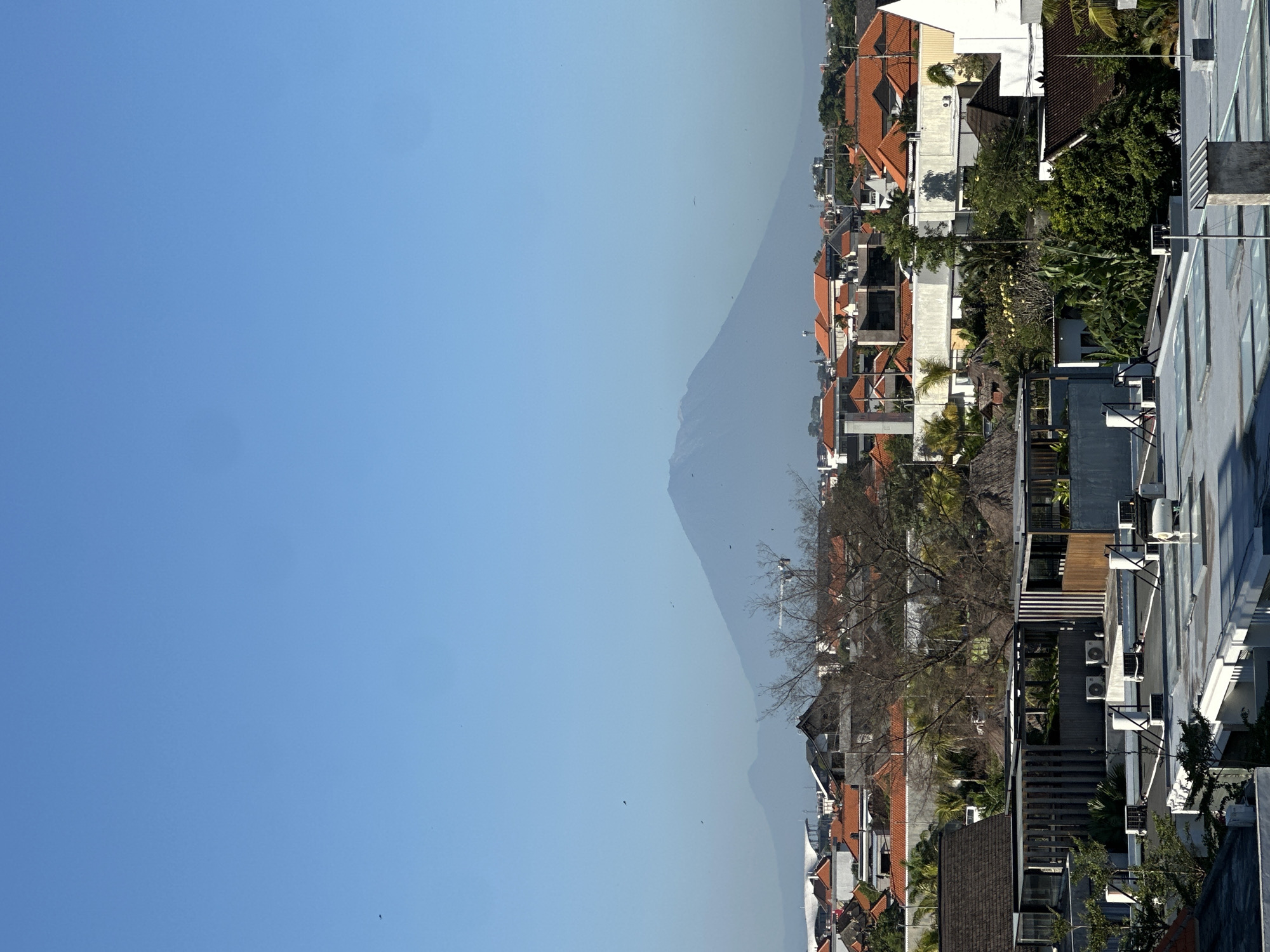 The image depicts a view in North Kuta, Indonesia, facing a mountain, possibly Mount Agung, in the distance. The right side of the image shows buildings of varying heights, most with terracotta-colored roofs. Air conditioning units are visible on the sides of some buildings, and trees are interspersed among the structures. The left side of the image is dominated by a clear, blue sky. Several small birds are visible against the sky. The mountain appears hazy in the background. The image is rotated 90 degrees clockwise.