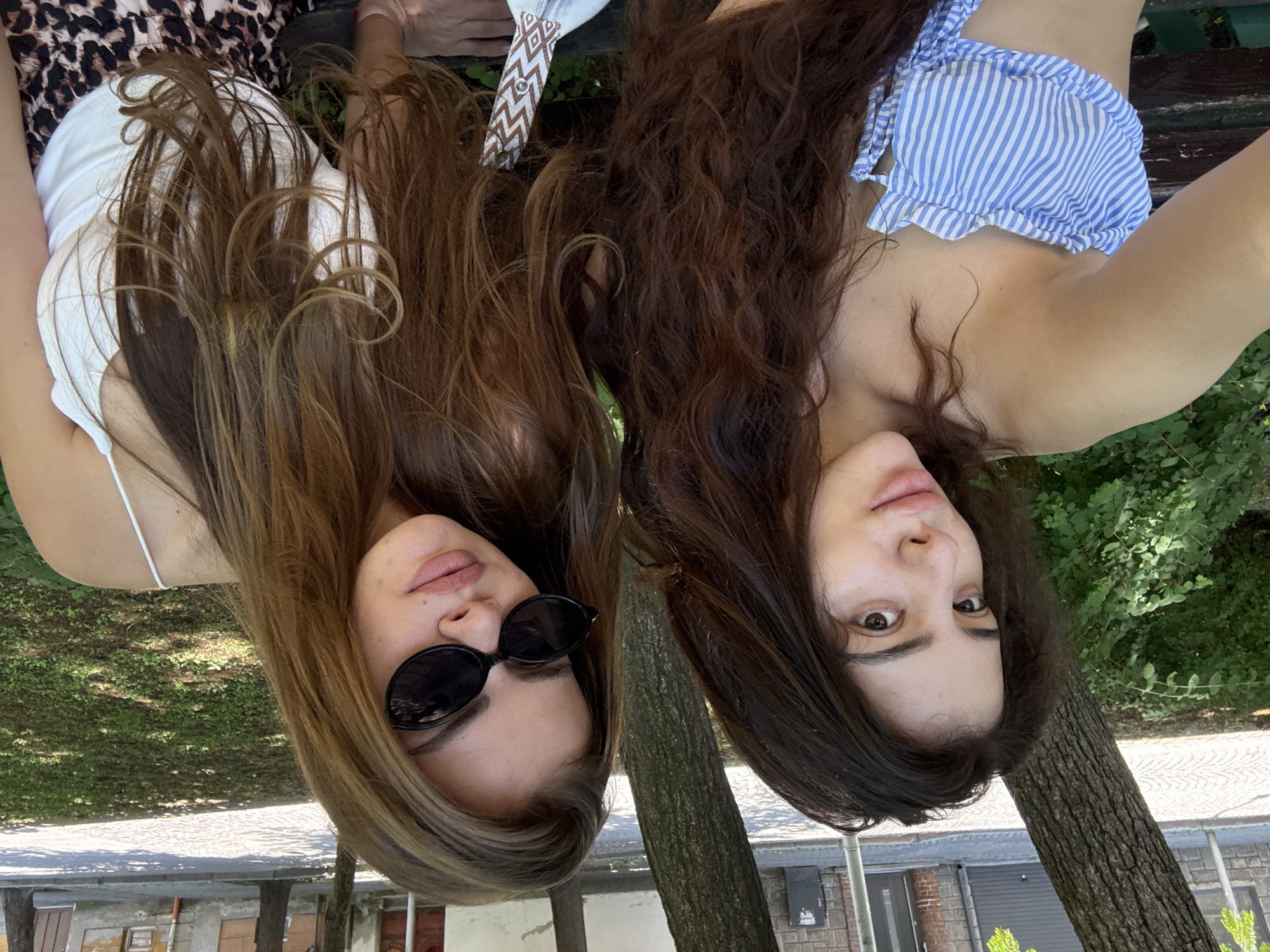 Two young women are seated outdoors on a park bench, taking a selfie. The woman on the left has long, dark brown, wavy hair and is wearing a blue and white striped off-the-shoulder top. The woman on the right has long, light brown, wavy hair, is wearing sunglasses and a white tank top, and has on leopard print pants. They are positioned in front of trees and lush green foliage, with a cobblestone path and buildings visible in the background. The lighting suggests it is daytime. The scene appears to be a public park within the city of Plovdiv, Bulgaria, likely near the Old Town given the architectural style of the buildings in the background and the presence of cobblestone streets.