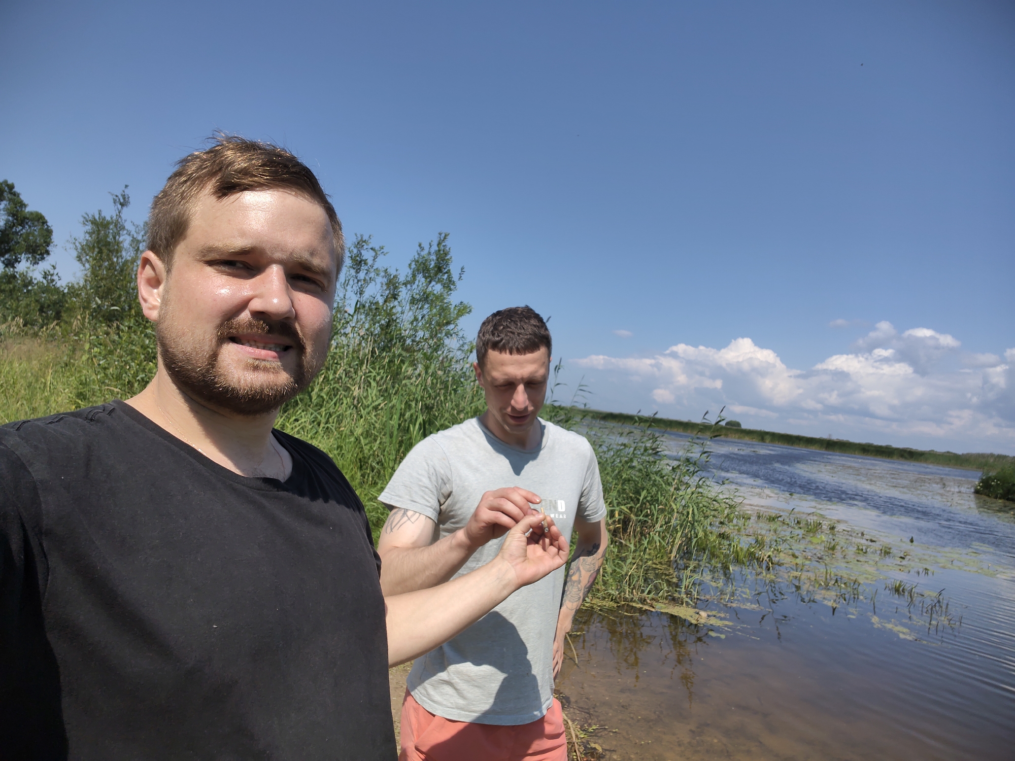 Two men stand outdoors on a sunny day with a body of water and reeds in the background. The man in the foreground, on the left, is wearing a black t-shirt and is looking towards the camera. He has light brown hair and a beard. The man in the background, on the right, is wearing a grey t-shirt and salmon-colored shorts. He is looking down at something he is holding between his fingers, which appears to be a cigarette. He has short brown hair and tattoos on his arms. The environment appears to be a rural area with a river or large pond, surrounded by tall grasses and trees. The sky is blue with scattered white clouds. The location is identified as Malaya Listovka, Russia.