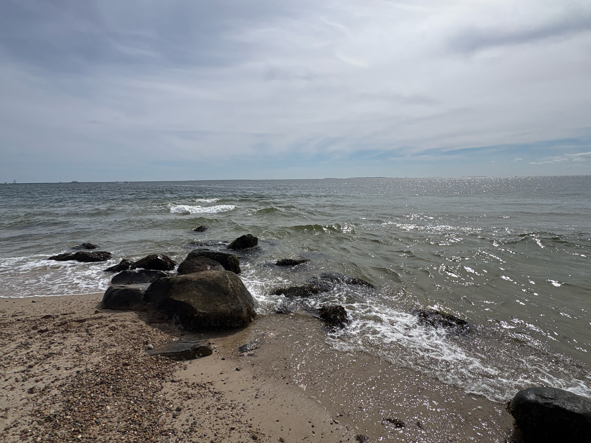 The image depicts a rocky shoreline on a cloudy day with a gentle surf. In the foreground, there is a sandy beach covered with small pebbles and several large, dark rocks. The water is a dark gray-blue, with small waves breaking against the shore, creating white foam. The horizon is visible in the distance, where the water meets a light gray sky with scattered white clouds. A few distant sailboats and ships can be seen on the water. The scene appears to be a natural coastal environment, possibly a public beach or park in Waterford, United States of America. There are no people or animals visible in the image.