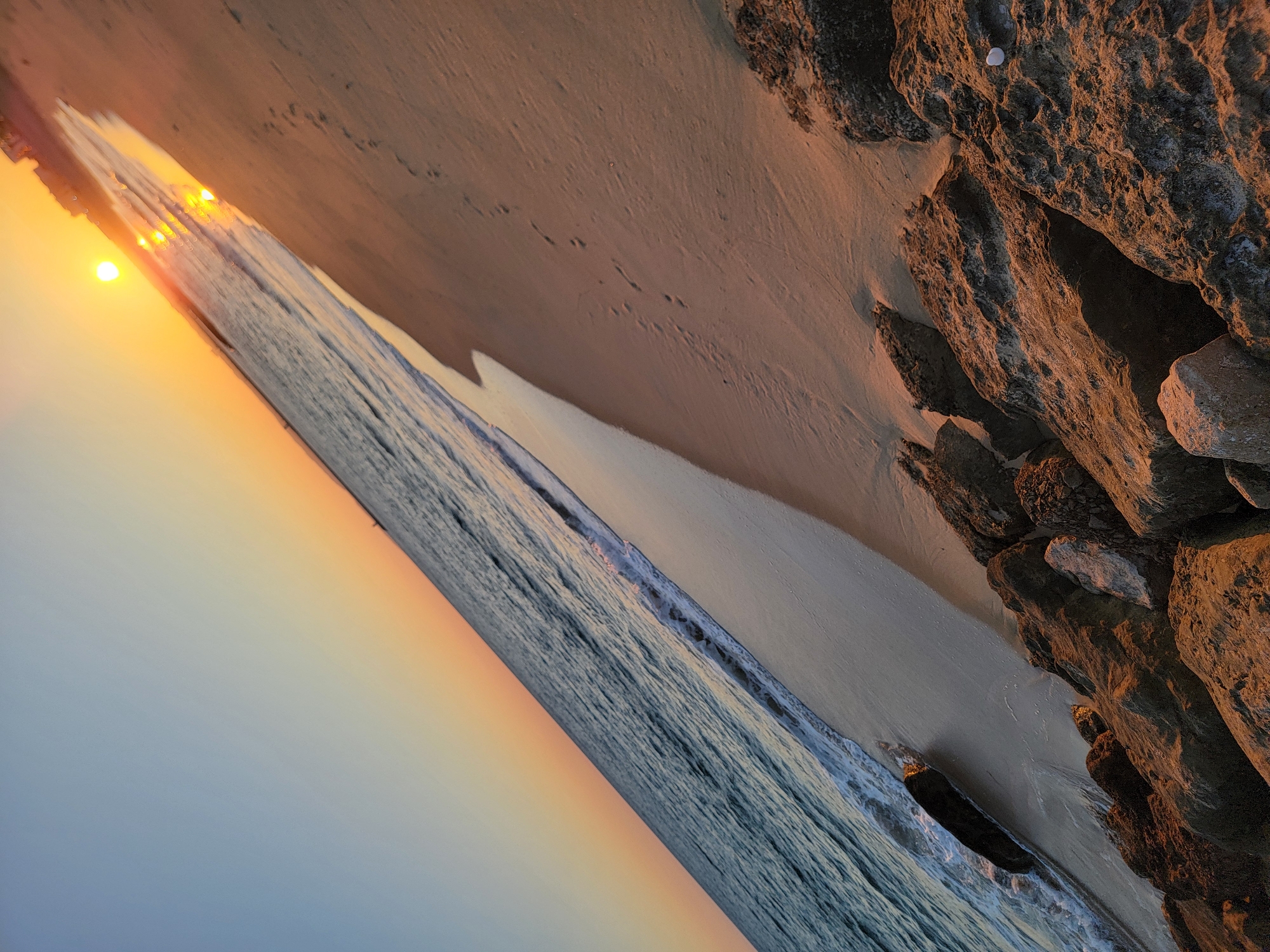 The image depicts a beach scene at sunset, likely in Quarteira, Portugal, given the tilt of the horizon and the presence of a sunset over the ocean. The sun is low in the sky, casting a warm, golden light across the sand and water. The ocean is a deep blue with gentle waves breaking on the shore. The sand is light brown and appears damp near the water's edge, with footprints visible further up the beach. There are rocks and boulders scattered along the shoreline, illuminated by the setting sun. In the distance, a faint outline of buildings or structures can be seen along the coast. The sky transitions from a pale blue at the top to a soft orange and yellow near the horizon.