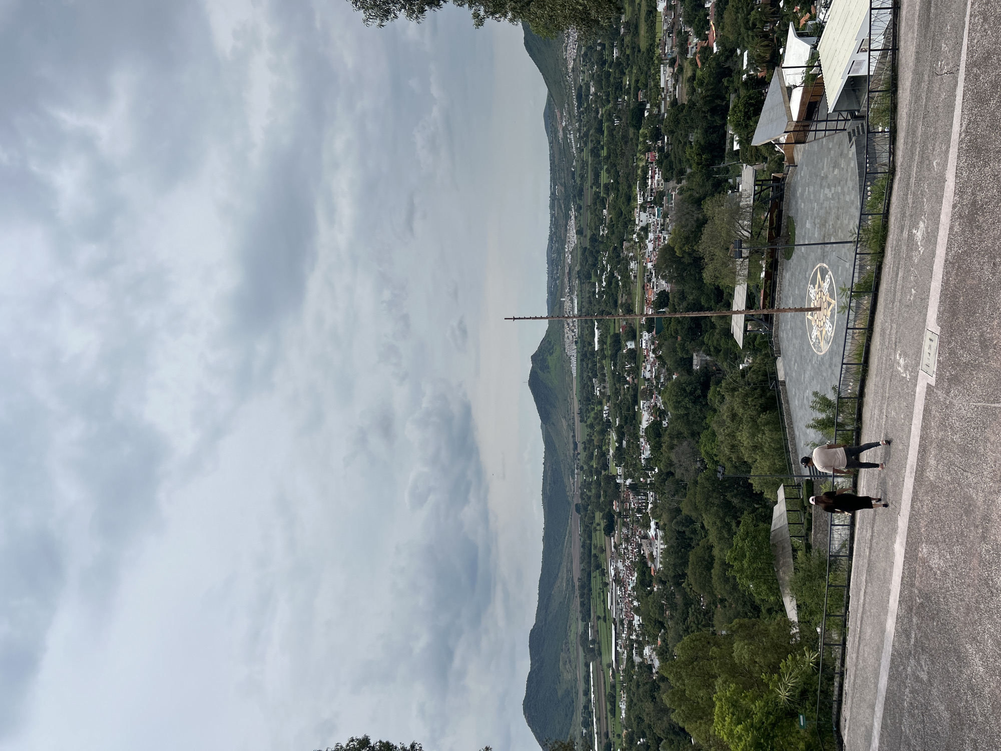 A high-angle, full shot captures a panoramic view of Atlixco, Mexico, under a cloudy sky. In the foreground, two individuals, a woman wearing a black dress and a pink cap, and a man in a light-colored shirt and dark pants, walk away from the camera down a paved slope. Beyond them, a circular plaza with a decorative mosaic on the ground is visible, featuring a flagpole at its center. The plaza is surrounded by a dark metal fence and is bordered by trees and modern architectural structures with white roofs. The midground reveals a sprawling town with red-tiled roofs and white buildings nestled amongst lush green trees. In the distance, rolling green hills form the backdrop of the scene.