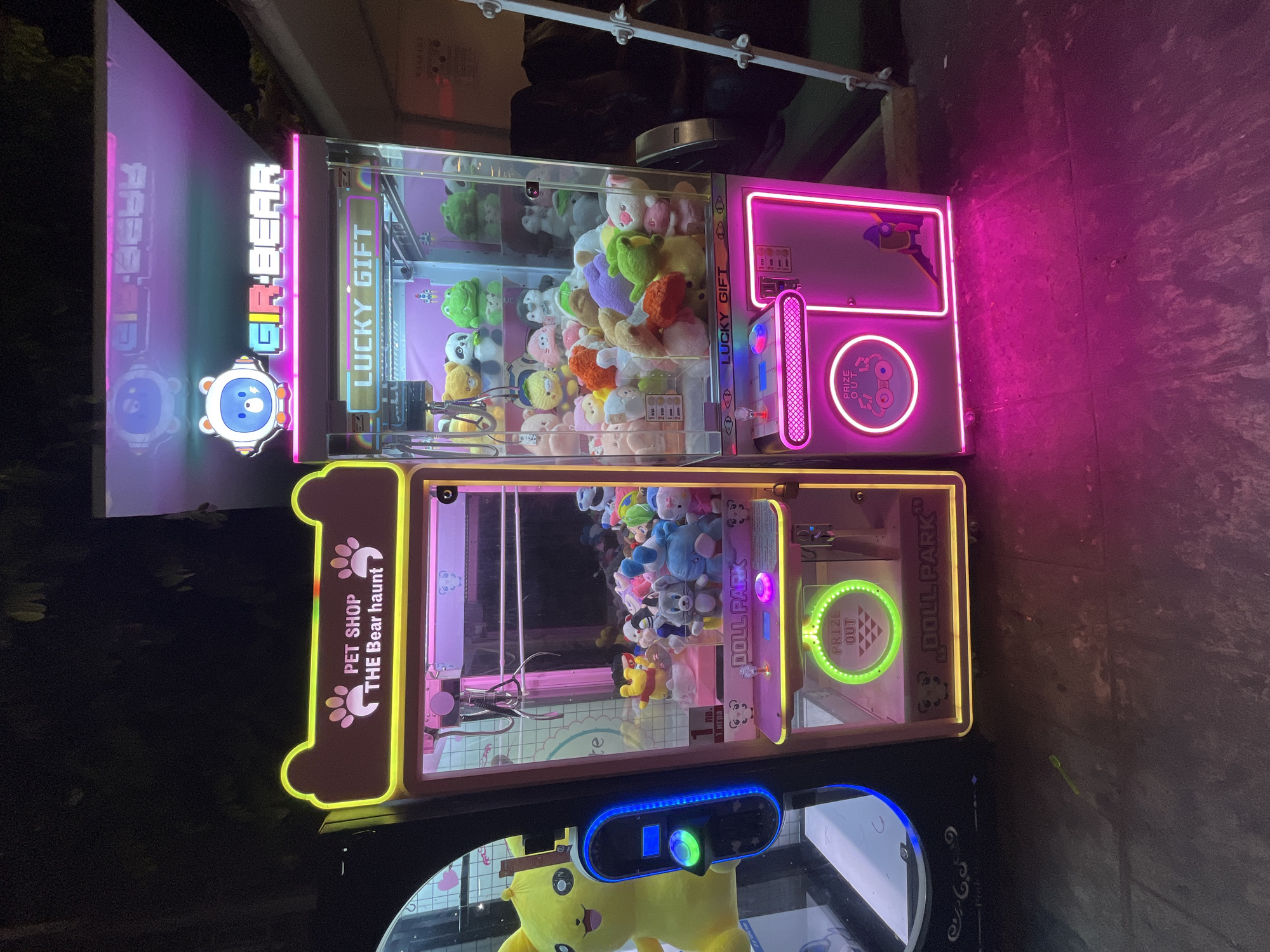Two claw machines are positioned next to each other outdoors at night. The machine on the left, labeled "PET SHOP THE Bear haunt," is primarily lit with yellow and pink neon lights. It contains a variety of plush toys, and its controls are illuminated with blue neon. To the right of this machine is a second claw machine, labeled "GR BEAR LUCKY GIFT," predominantly lit with pink neon lights. This machine also houses numerous plush toys, including animals like pandas and frogs. The ground in front of the machines is paved, and the background suggests an urban setting with some foliage and structures. A tall, white ladder-like structure is visible to the right of the second claw machine. No people are visible in the frame. The location is identified as Obzor, Bulgaria.