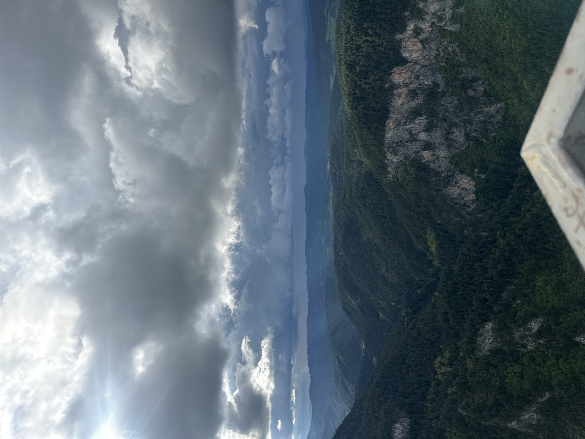 The image depicts a view from an elevated position overlooking a mountainous landscape, likely near Iagodina, Bulgaria. The foreground features a corner of a structure, possibly a viewing platform. The landscape consists of a series of forested ridges receding into the distance, with exposed rocky cliffs visible on some slopes. Weather conditions appear overcast; heavy clouds fill the sky, with some areas showing brighter patches of light. The horizon is partially obscured by haze and overlapping mountain ranges.