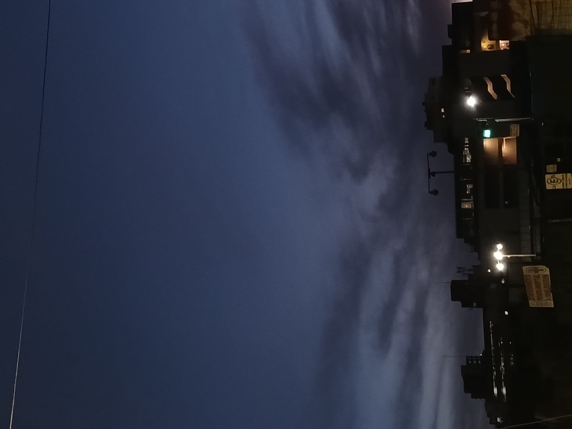The image captures a nighttime scene in Buenos Aires, Argentina. The right side of the frame shows a dark urban environment, with buildings, streetlights, and signs silhouetted against the dusky sky. The left side of the image is dominated by the dark blue sky, with streaks of clouds adding texture.