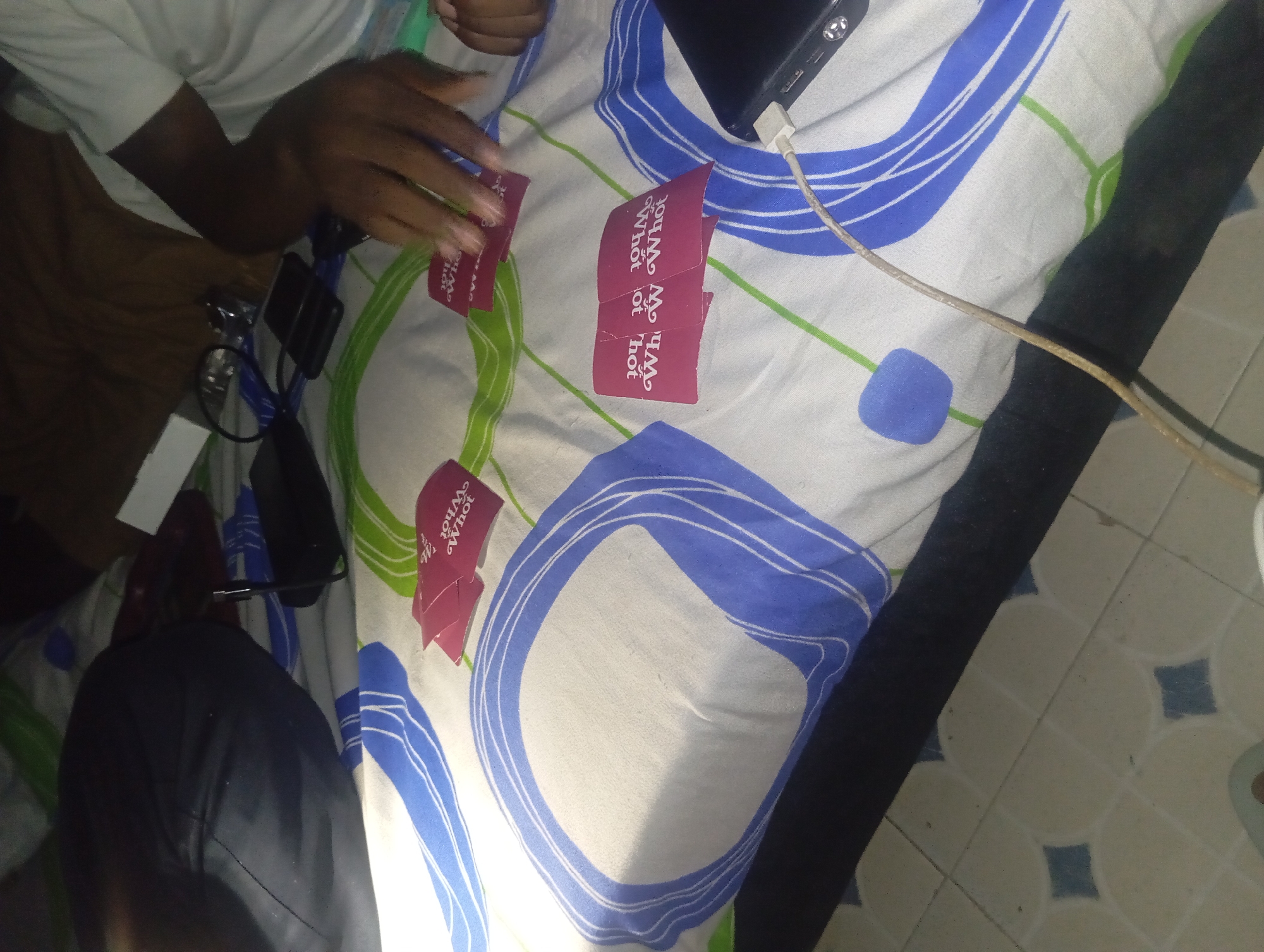 The image depicts a close-up, overhead view of a person's hands interacting with red playing cards on a white and blue patterned surface. The cards have white text that reads "You, or What". The person's hands are dark-skinned and appear to be holding and shuffling the cards. A black smartphone is visible in the lower right corner, plugged into a charger, with a white cable running across the surface. The background is a cluttered bed with a dark pillow or bag to the left. The environment suggests an indoor setting, possibly a bedroom or living area. The overall lighting is dim.