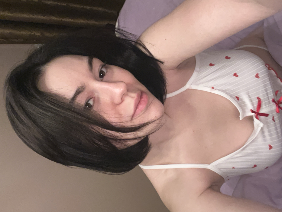 A woman with short, dark hair lies on a purple surface, facing the camera. She wears a white top decorated with small red hearts and a red ribbon accent. Her expression is serene, and her skin appears clear. The lighting is soft, creating a warm tone across the image. The background is simple, with a hint of drapery visible in the upper left corner. The angle is slightly tilted, giving a close-up view of her face and upper body.