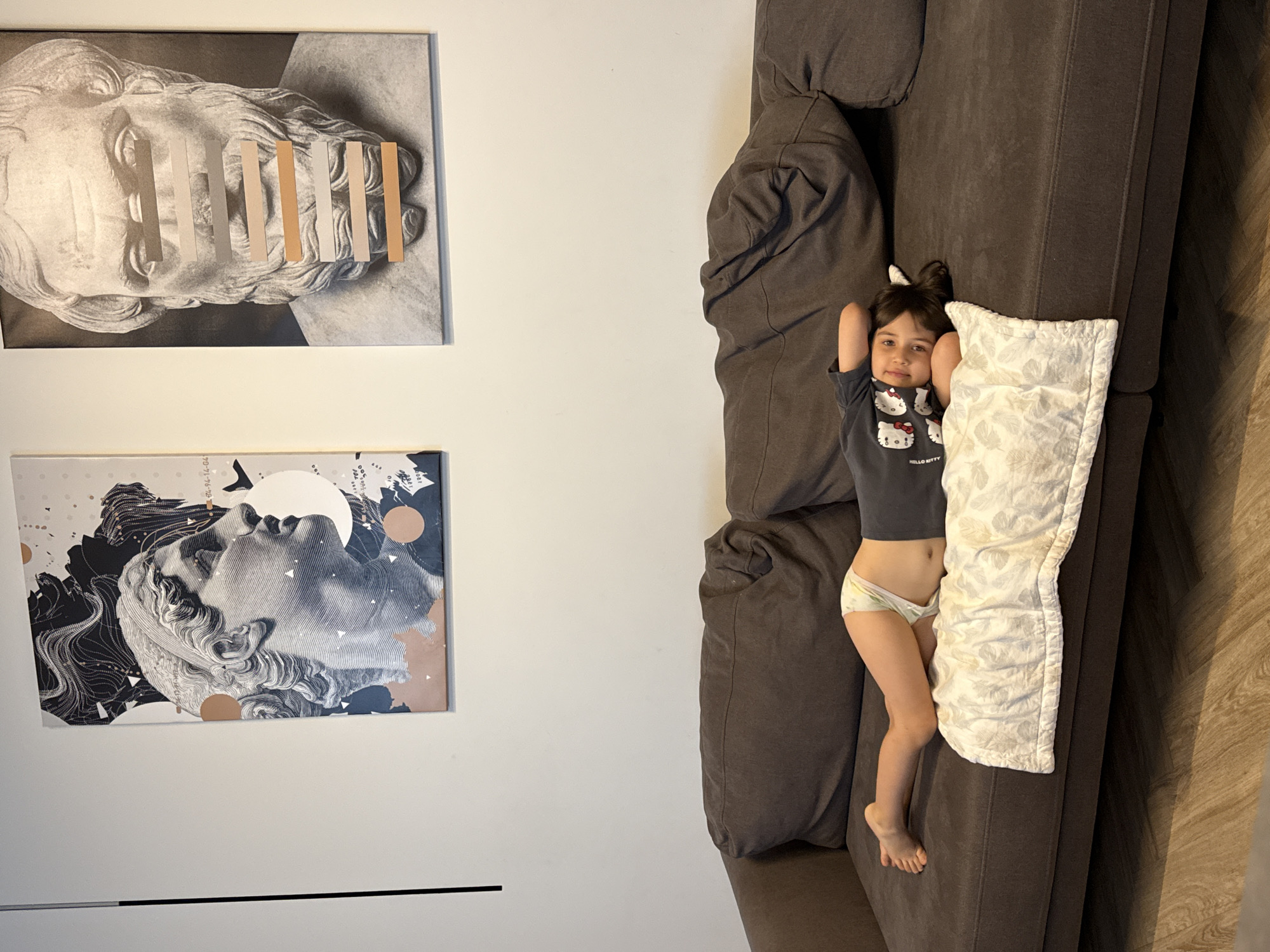 A young girl is lying on a brown couch with a pillow behind her head. She is wearing a gray shirt with cartoon characters and white underwear. Her hands are behind her head. Two framed pieces of art are on the wall. One depicts a black and white sculpture with colored lines over the eyes. The other is a black and white illustration of a sculpture with abstract designs. The couch is positioned in a room, likely a living room, next to a light-colored wooden floor. The photograph was taken indoors, and there is no indication that it is related to Krasnoyarsk, Russia.