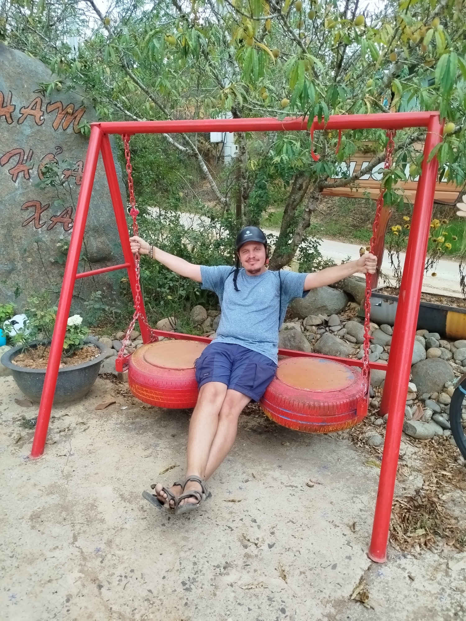 In Đa Thọ, Vietnam, a man is seated on a tire swing within a park-like setting. He is wearing a gray shirt, blue shorts, sandals, and a helmet. The swing is painted red, and the seats are made of repurposed tires. Surrounding the swing are rocks, plants, and a large stone with text carved into it.