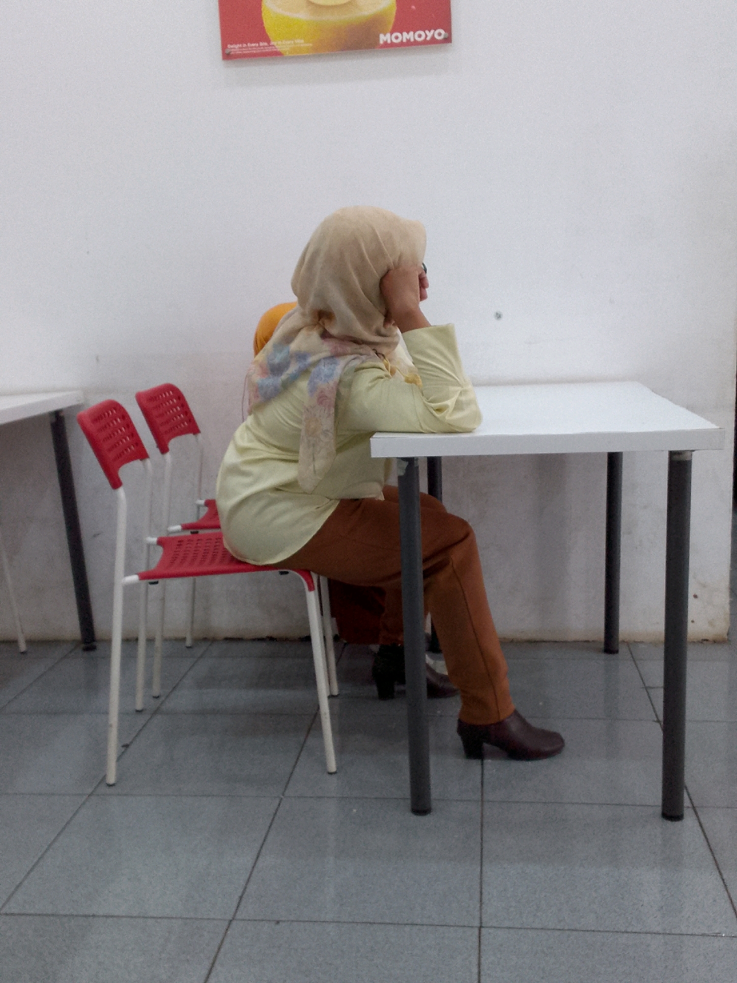 A woman wearing a tan hijab and a light yellow long-sleeved shirt sits at a white rectangular table, her right arm resting on the table and her left hand touching her head. She wears brown pants and dark brown heeled boots. In front of her, three red plastic chairs with white metal legs are visible. The room has a white wall and a grey tiled floor. A sign with the word "MOMOYO" is partially visible on the wall behind the woman. The scene appears to be indoors, possibly a restaurant or waiting area.