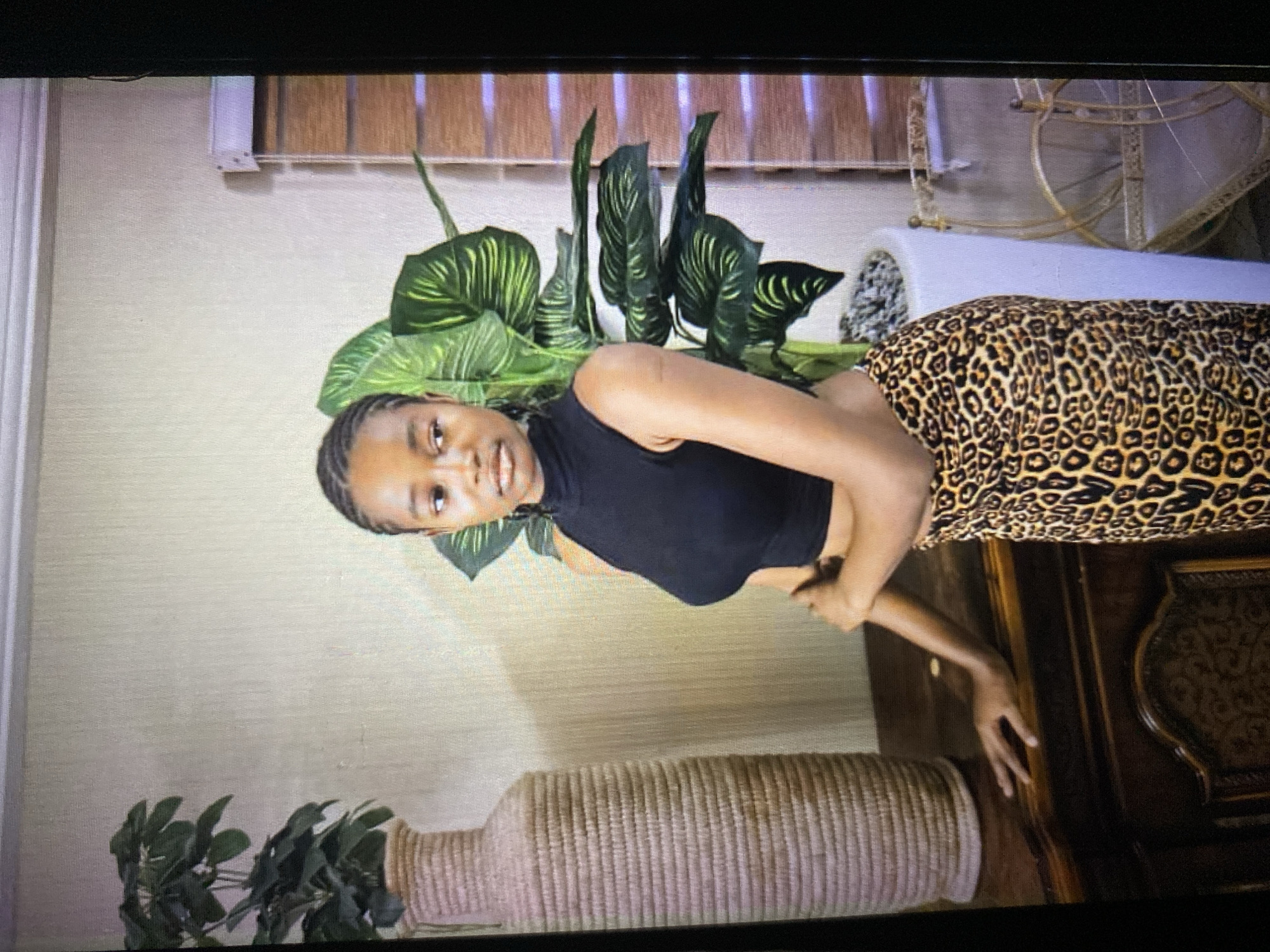A young woman with dark braided hair stands indoors, leaning against a dark wooden dresser. She is wearing a black, sleeveless, high-neck crop top and a long leopard-print skirt. To her left is a tall, textured vase with a green plant. Behind her and to her right is a large potted plant with dark green leaves that have yellow-green streaks. The wall behind the woman appears to be a textured beige. To the far right, a wooden blind is partially visible, and a gold decorative chair or stand.