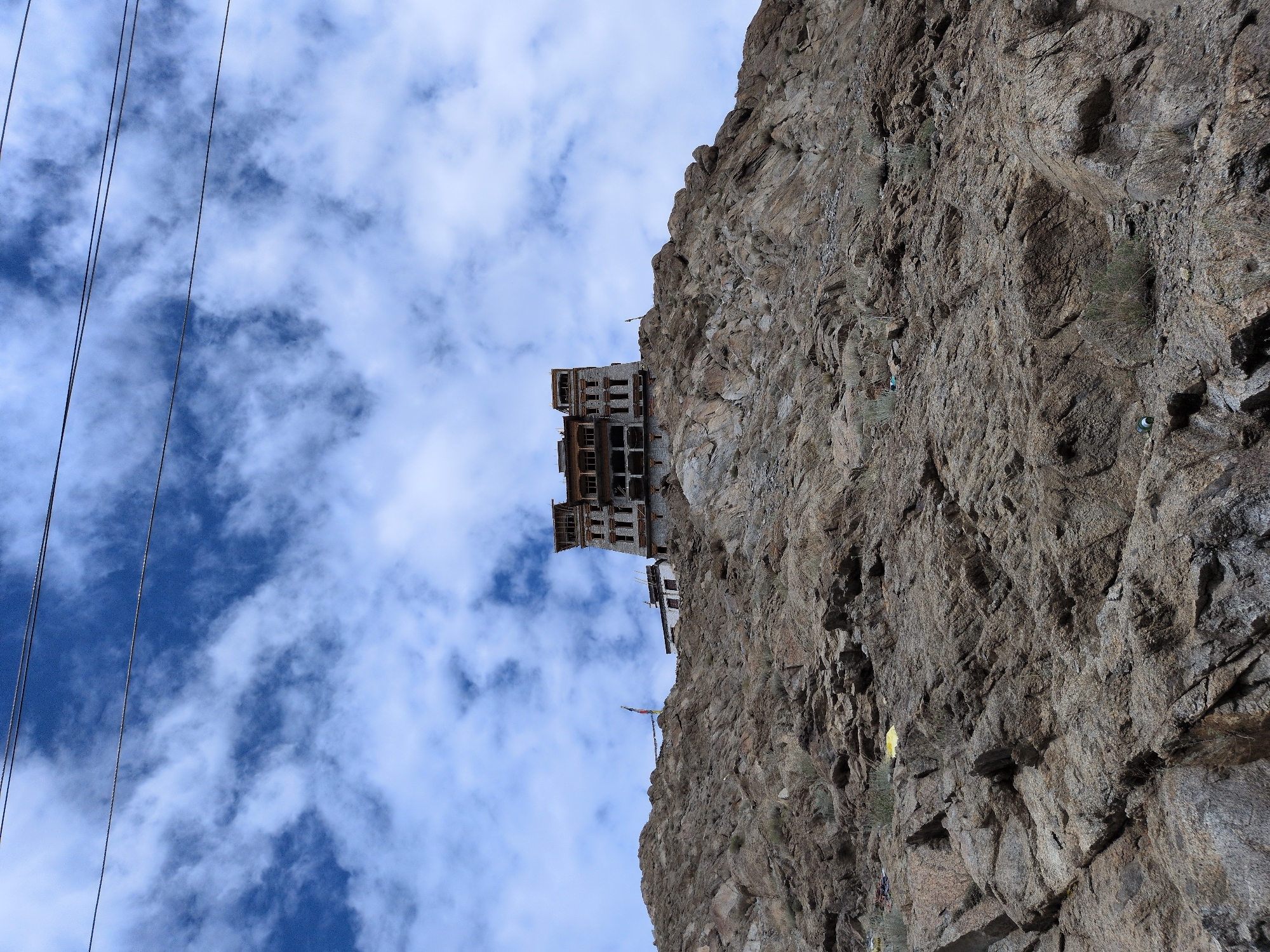 The image depicts a monastic complex perched atop a rugged, rocky mountain. The architecture of the monastery is traditional, with multiple stories and intricate stonework. Prayer flags flutter on a pole to the left of the main structure, adding a touch of color to the scene. Below the monastery, the mountainside is barren and rocky, with sparse vegetation. The sky above is a vibrant blue, dotted with white clouds, and two power lines stretch across the upper portion of the frame. This is likely the Spituk Monastery in Leh, India, a prominent Buddhist monastery situated on a hilltop overlooking the Indus River.