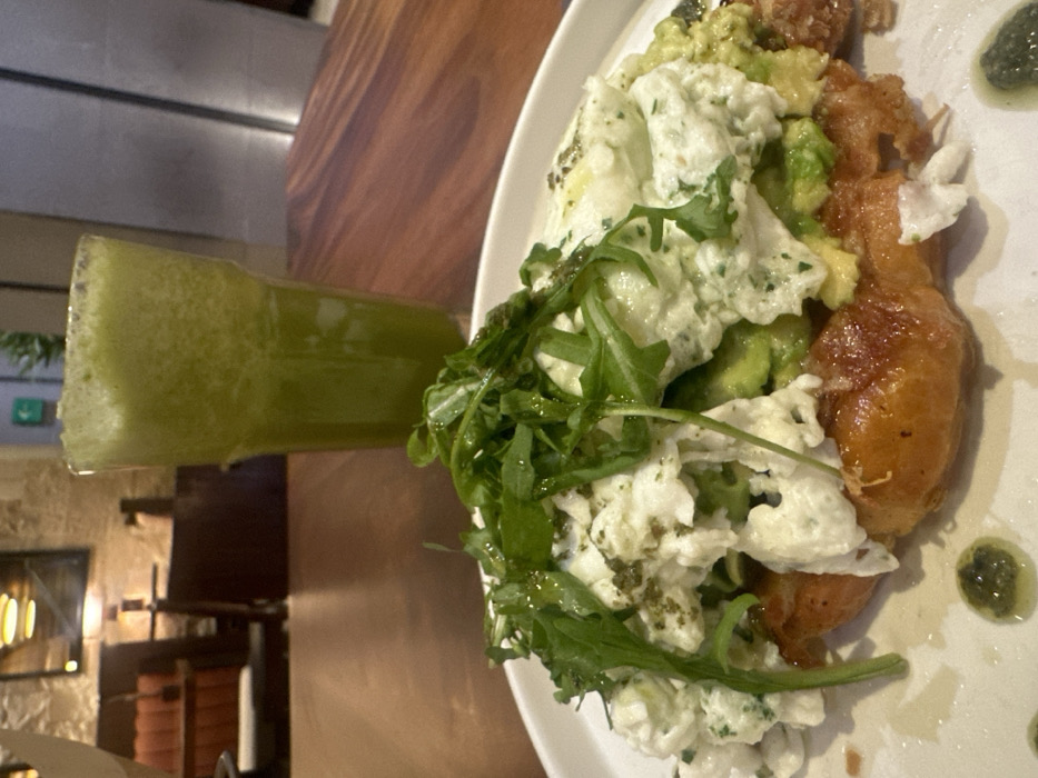This image depicts a meal served on a wooden table within a restaurant in Mexico City, Mexico. In the foreground, a white oval plate holds a dish featuring a golden-brown pastry base topped with mashed avocado, a substantial portion of scrambled eggs or ricotta cheese mixed with herbs, and a generous covering of fresh arugula leaves. Small droplets of green herb oil are scattered on the plate. To the right and slightly behind the plate, a tall, clear glass contains a vibrant green, foamy beverage. The background, out of focus, reveals the restaurant's interior: a textured stone wall on the left, dark upholstered seating, warm-toned lighting fixtures, and a framed item. On the right side of the background, modern, dark wall panels are visible, and a green illuminated exit sign is faintly discernible in the upper left. No individuals are present in the scene.