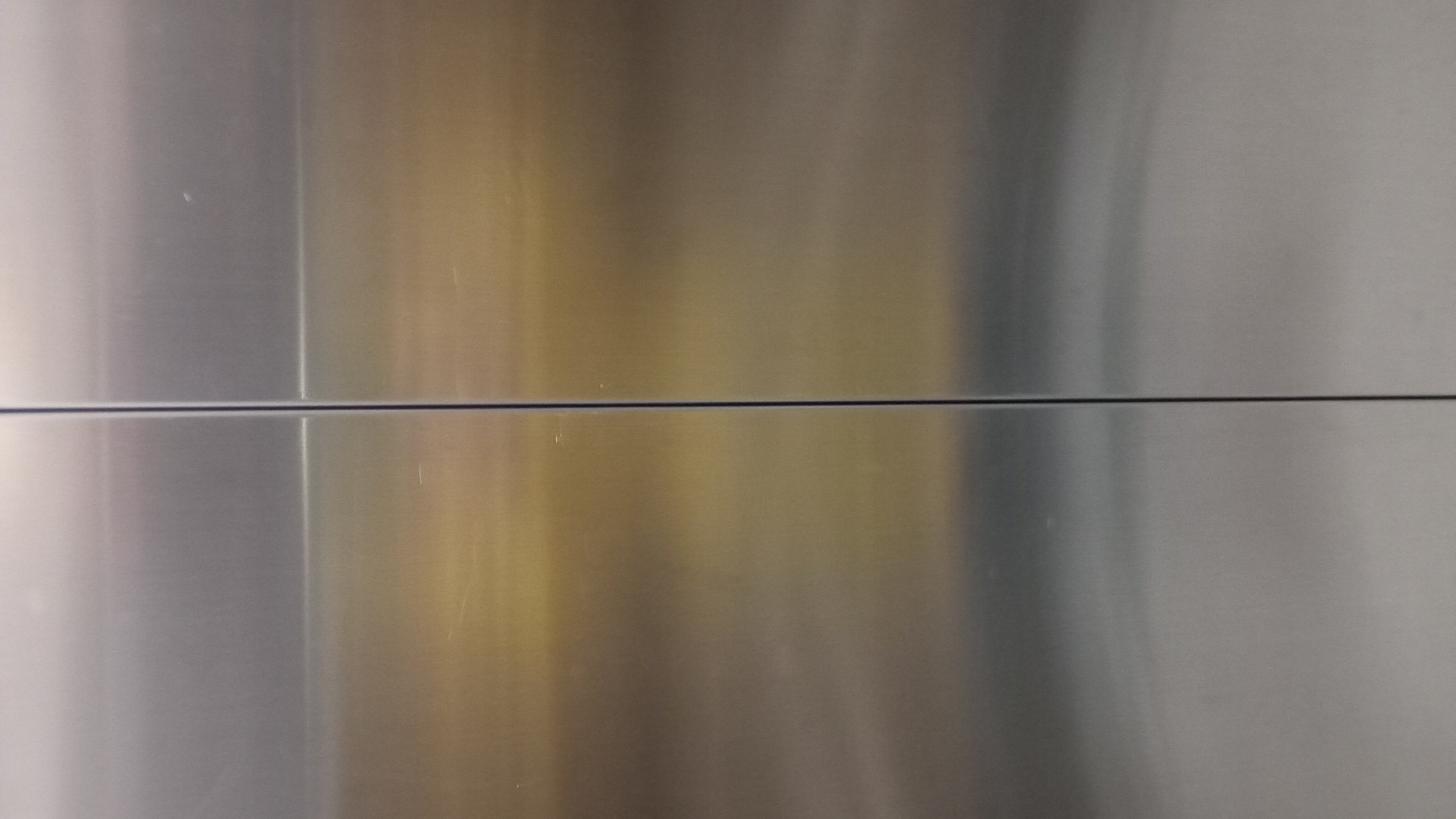 The image shows a close-up of a metal surface with a horizontal seam. The metal has variations in color, appearing silver, gold, and grey. There are minor scratches on the surface. The location and connection to Batumi, Georgia, are not apparent.