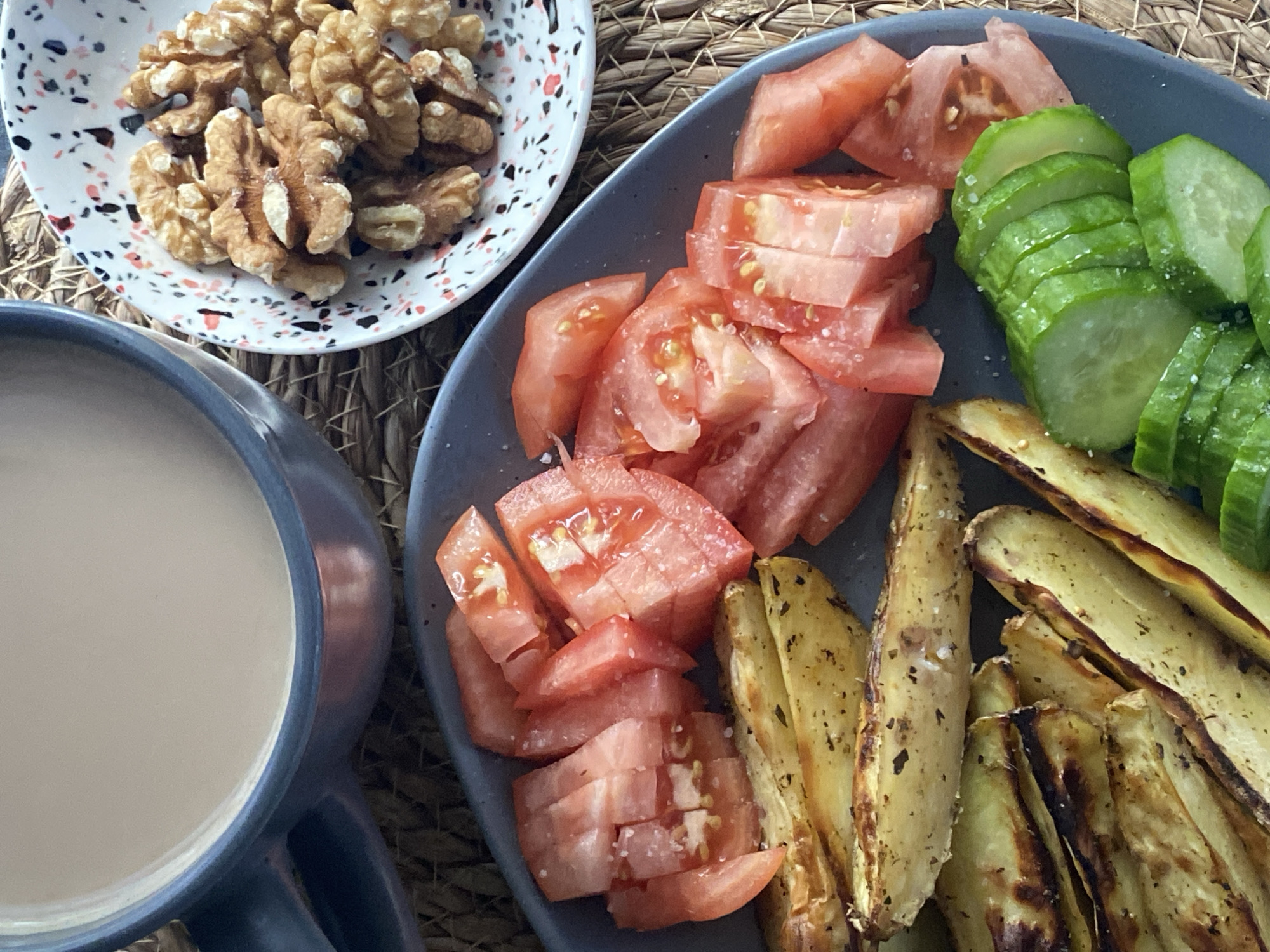 The image shows a close-up, overhead view of a breakfast spread. In the foreground, a dark blue plate holds several food items. On the left are baked potato wedges, seasoned with herbs and salt, with some appearing slightly browned. To the right of the wedges are chopped tomatoes, also seasoned with salt, and sliced cucumbers sprinkled with salt. In the background, a blue mug filled with a light-colored beverage, likely coffee or tea with milk, sits on a woven placemat. To the right of the mug, a small white bowl with a speckled pattern contains a pile of walnut halves. The scene is illuminated by natural light, casting soft shadows. There are no people or animals visible in the image. The environment appears to be an indoor setting, possibly a dining table. The image does not contain any information that would allow for its location to be identified in the city of Chaikovsky, Russia.