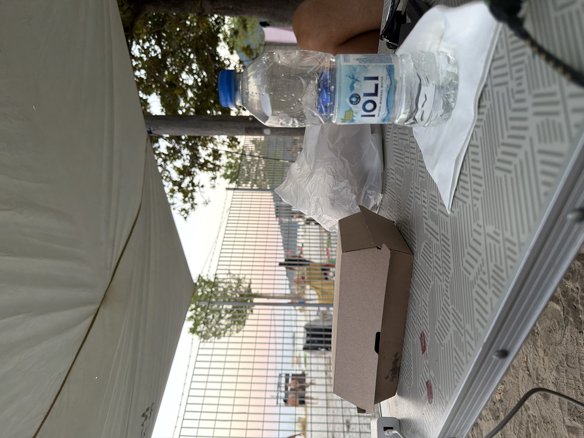 The image depicts a table set up outdoors, likely on a beach, with a partially consumed bottle of IOLI natural mineral water and a takeaway food container. The table is positioned under a white canopy, offering shade from the sun. In the background, a metal fence separates the seating area from a beach with people enjoying the water. The sky shows a soft gradient of colors, suggesting it might be sunset or sunrise. The overall scene conveys a sense of casual outdoor dining in a coastal environment, potentially within Golden Beach, Greece.