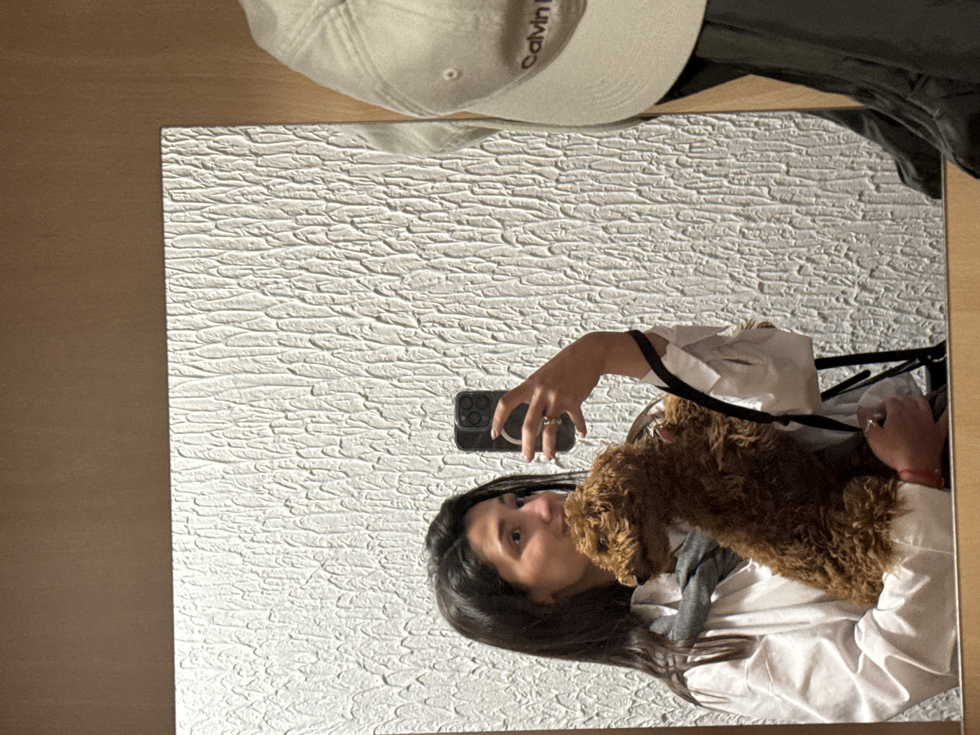 A mirror reflects a woman holding a small, brown dog while taking a selfie with her smartphone. A white Calvin Klein baseball cap and black clothing item are visible near the top edge of the mirror, presumably on a surface above. The mirror is in a room with a textured white wall, and a wooden surface is visible on the left side of the mirror. There is no evidence to confirm a location in Zankovići, Montenegro.