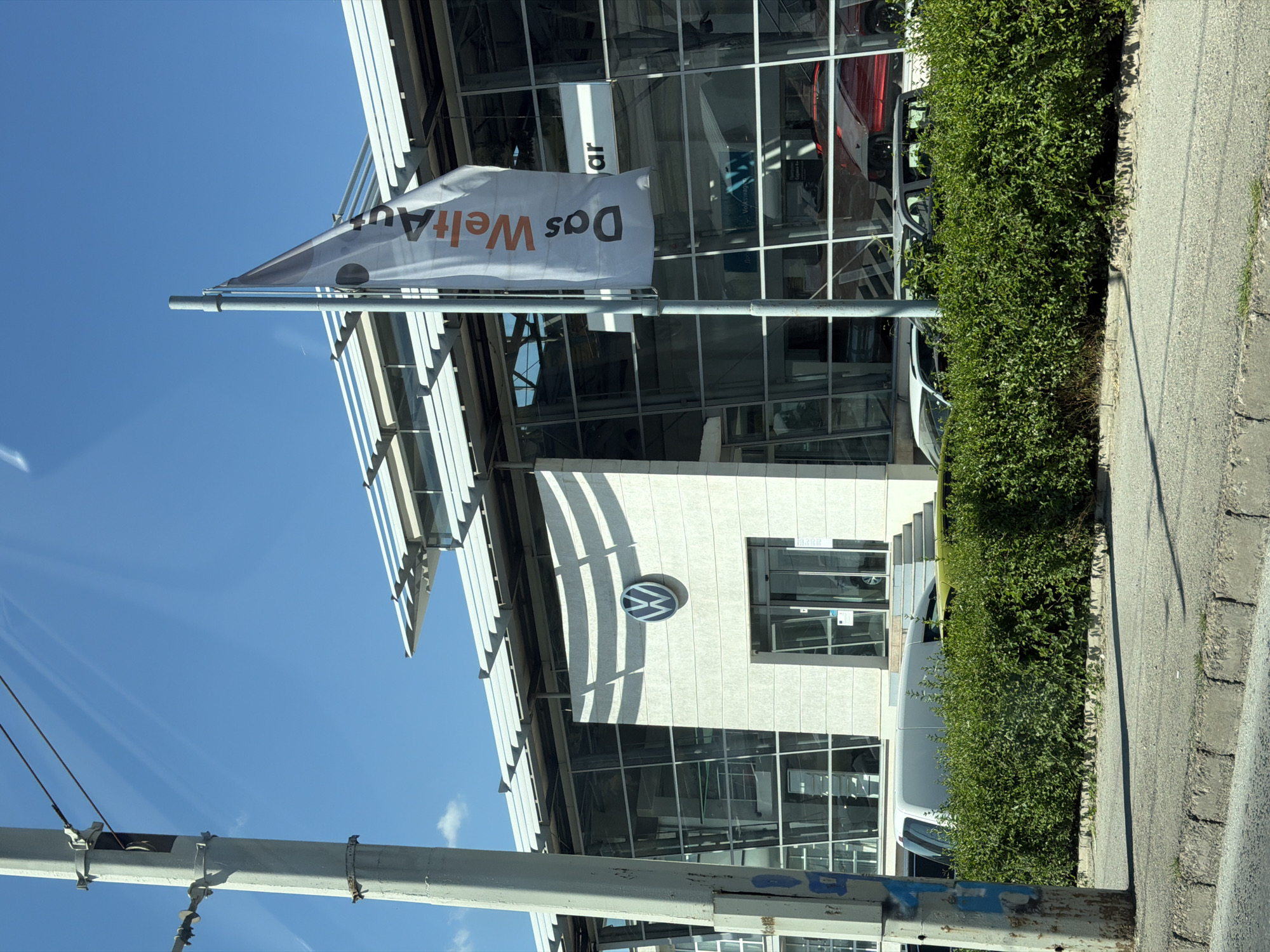 The image depicts the exterior of a Volkswagen dealership in Dolno Ablanovo, Bulgaria, under a clear blue sky. The building features a modern architectural design with extensive glass facades, a white stone-clad entrance area, and an overhanging roof structure. A prominent Volkswagen logo is visible on the facade above the entrance. A white flag with the text "Das WeltAuto" is displayed on a flagpole to the right of the entrance. In front of the building, a neatly trimmed green hedge separates the property from the sidewalk and road. Several vehicles are visible through the glass, including what appears to be a white van, a red sports car, and other vehicles. A utility pole stands on the left side of the image, partially obscuring the view of the building. The scene appears to be a standard day with no visible individuals engaged in any particular activity.