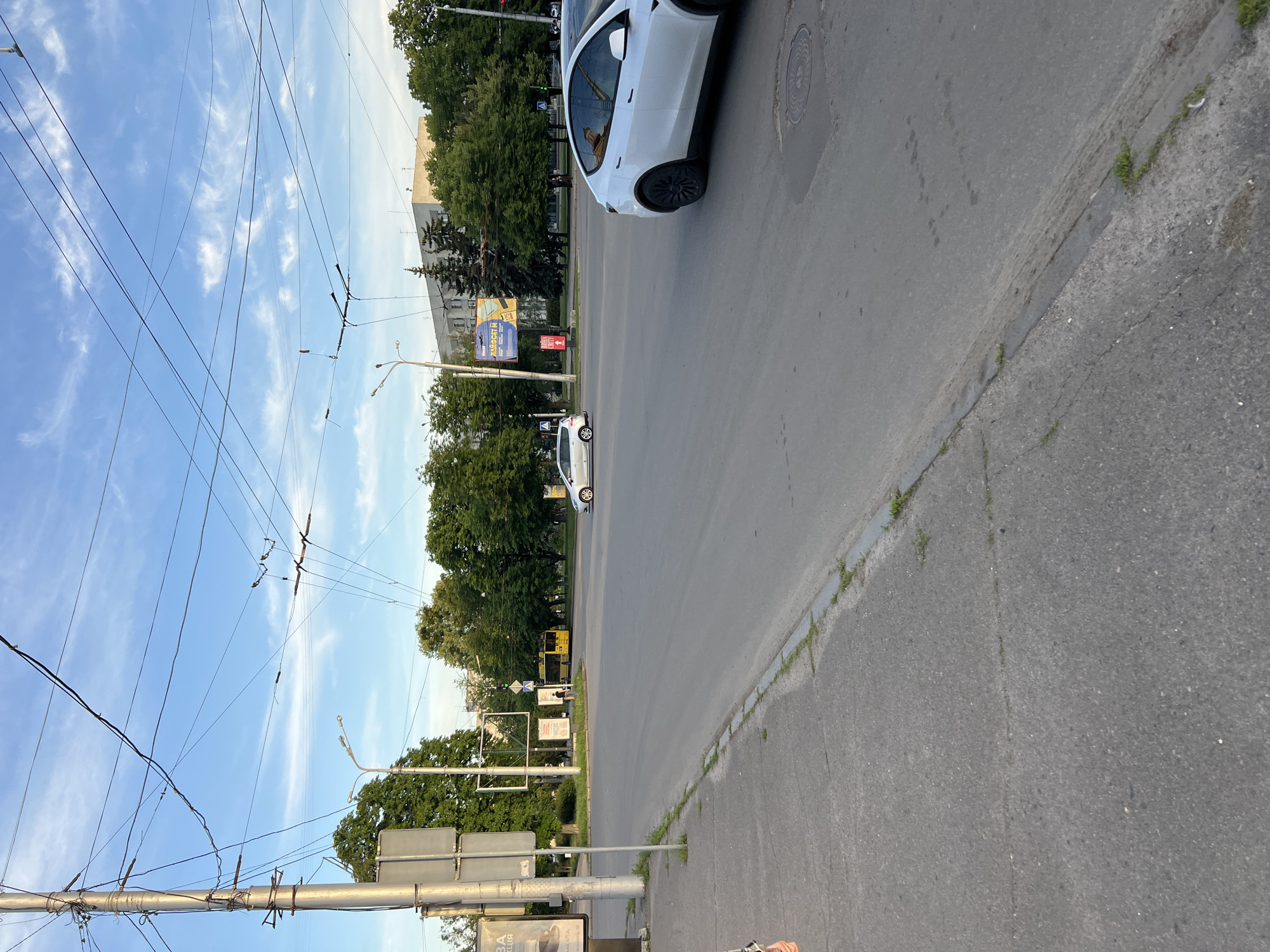 The image, taken in Poltava, Ukraine, captures a daytime urban street scene under a bright blue sky with scattered white, wispy clouds. A complex network of overhead utility and trolleybus wires spans across the upper portion of the frame.
The foreground is dominated by a paved asphalt road, with a curb and sidewalk visible along the right edge where small patches of grass grow. A white car, resembling a Tesla Model 3 or Y, is prominently positioned on the right side of the road, facing away from the viewer. Further down the street, another smaller white car is visible. A yellow public transport vehicle, likely a trolleybus or bus, can be seen in the distance, partially obscured by the dense green trees that line both sides of the thoroughfare.
Several utility poles support the overhead wires, and streetlights are also attached to these poles. Behind the trees, various buildings are discernible, contributing to the urban environment. A large blue and yellow billboard stands out, displaying Ukrainian text including "ПРАЦЮЙ" (Work), "ОДЯГ" (Clothing), and "ЗНИЖКИ" (Discounts). A smaller sign also features the word "АКЦІЯ" (Promotion/Sale). The scene portrays a typical, calm day with everyday traffic, characterized by the infrastructure for public transportation. The road shows some signs of wear, and a manhole cover is visible on its surface.